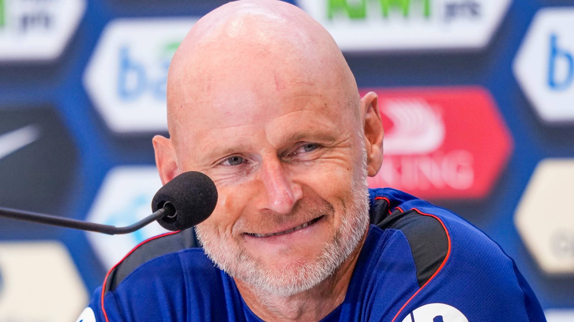 En mann sitter ved et podium og smiler. Han har kort, grått skjegg og er skallet. Bak ham er det en bakgrunn med logoer. Mannen har på seg en blå skjorte med en rund krage. (Bildebeskrivelsen er laget av en KI-tjeneste)