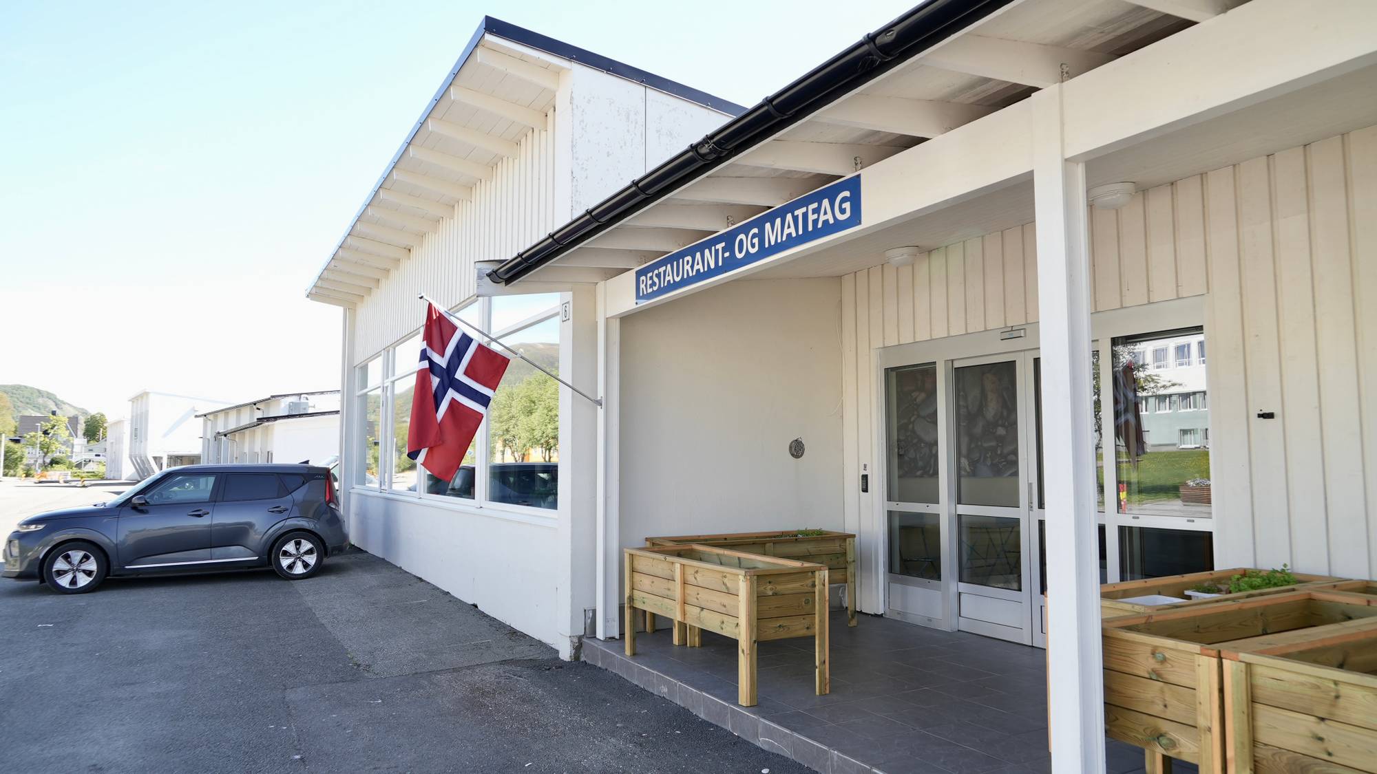 Et mørkegrått kjøretøy parkert utenfor en bygning med et skilt hvor det står "RESTAURANT - OG MATFAG". Et norsk flagg henger på bygningen. Utenfor inngangspartiet står det to blomsterkasser i tre. Solen skinner og himmelen er lys. (Bildebeskrivelsen er laget av en KI-tjeneste)