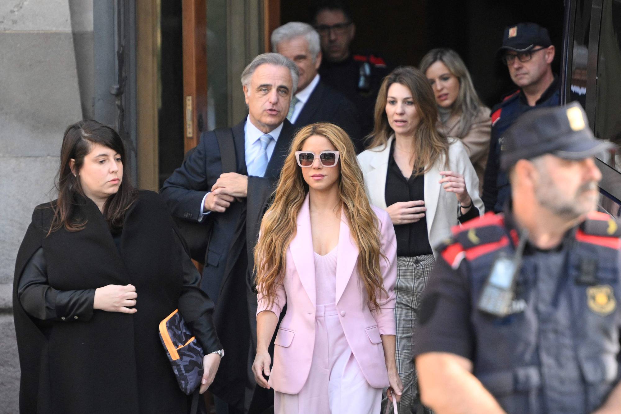 Artisten Shakira, iført lyserosa dress, forlater en bygning. Hun ledsages av menn og kvinner, samt politi. (Bildebeskrivelsen er laget av en KI-tjeneste)