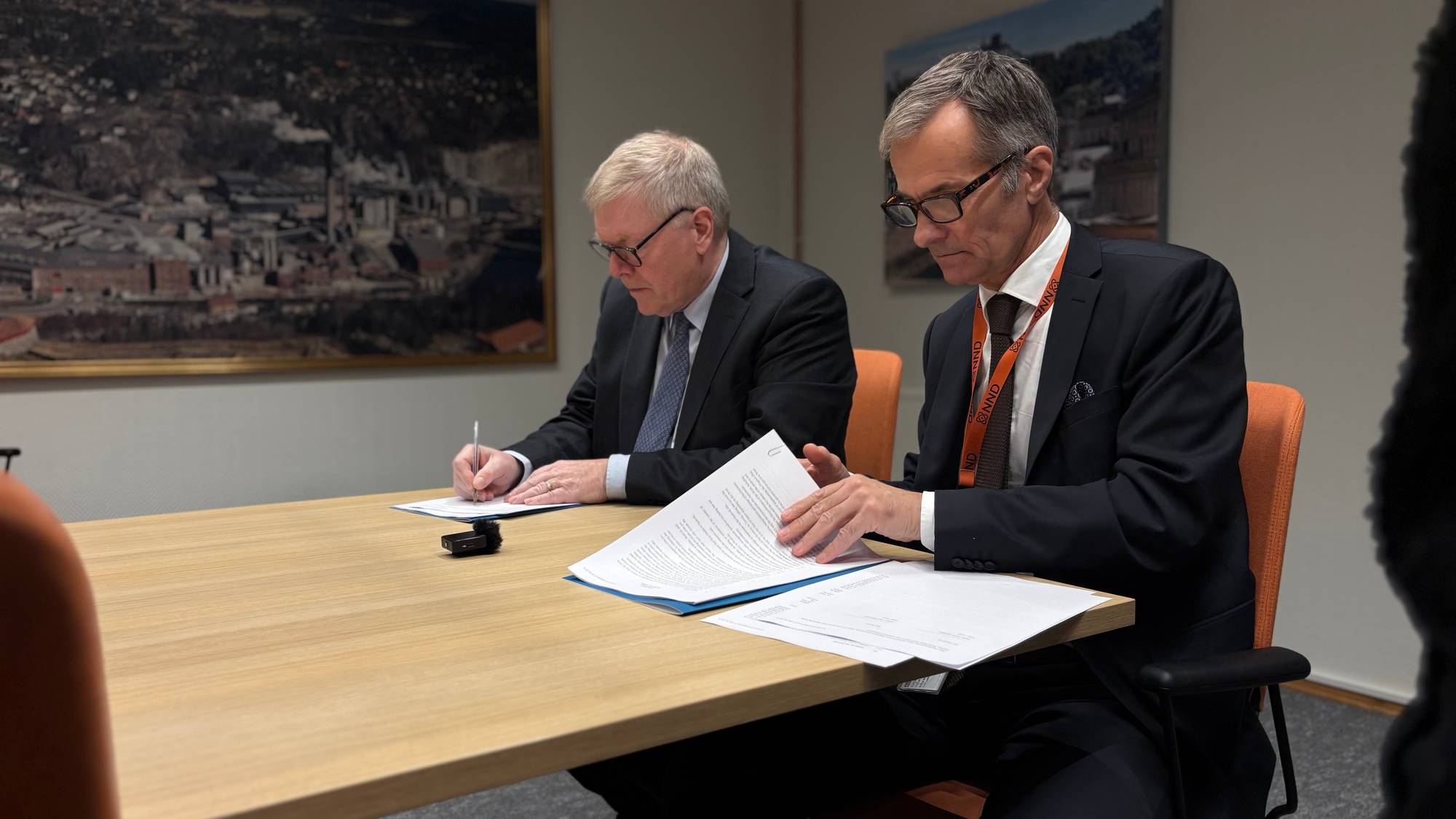 To menn i dress sitter ved et bord og signerer dokumenter i et møterom. (Bildebeskrivelsen er laget av en KI-tjeneste)