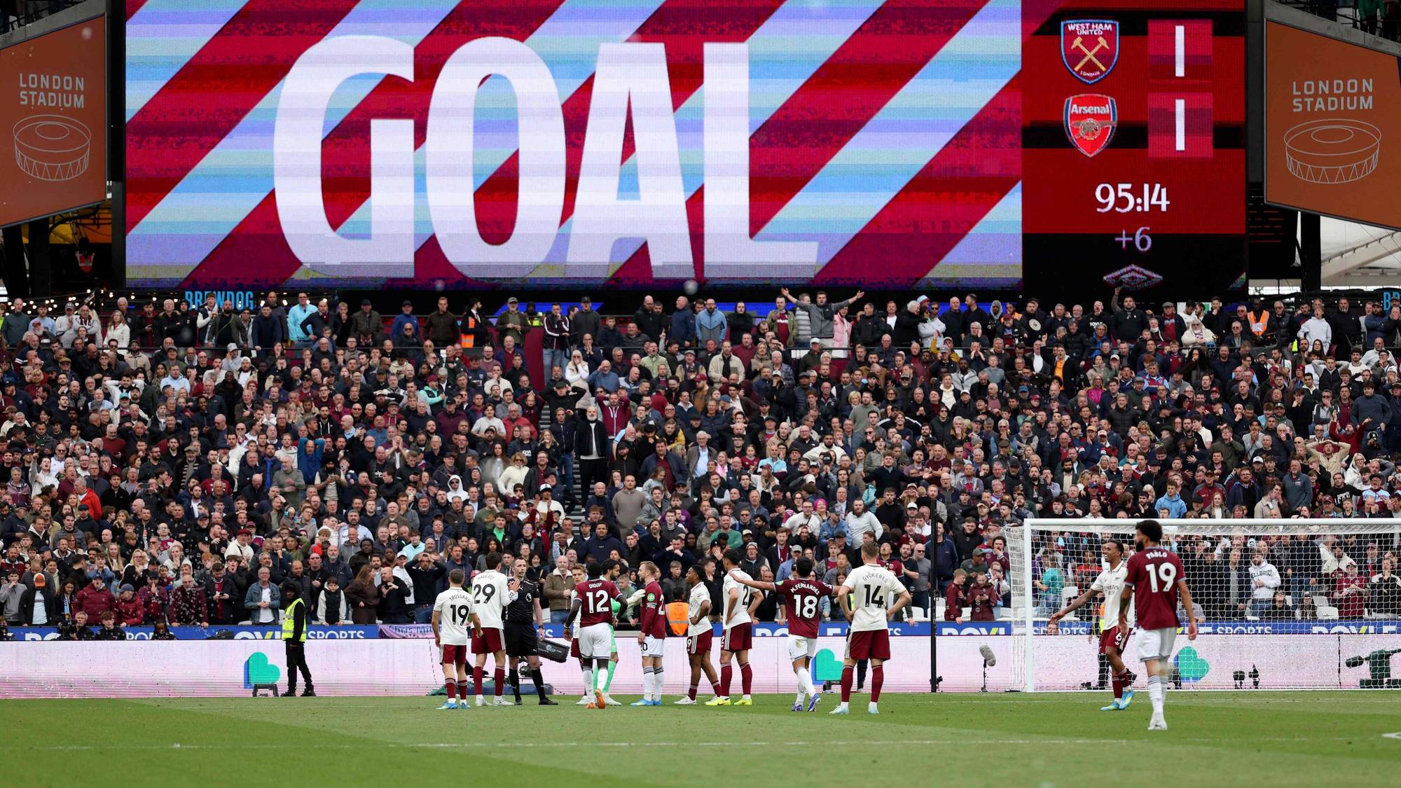 En stor skjerm viser ordet "GOAL" og resultattavlen for en fotballkamp. Et stort publikum fyller tribunene på et stadion. Spillere samles på gressmatten, noen av dem mot målet. (Bildebeskrivelsen er laget av en KI-tjeneste)