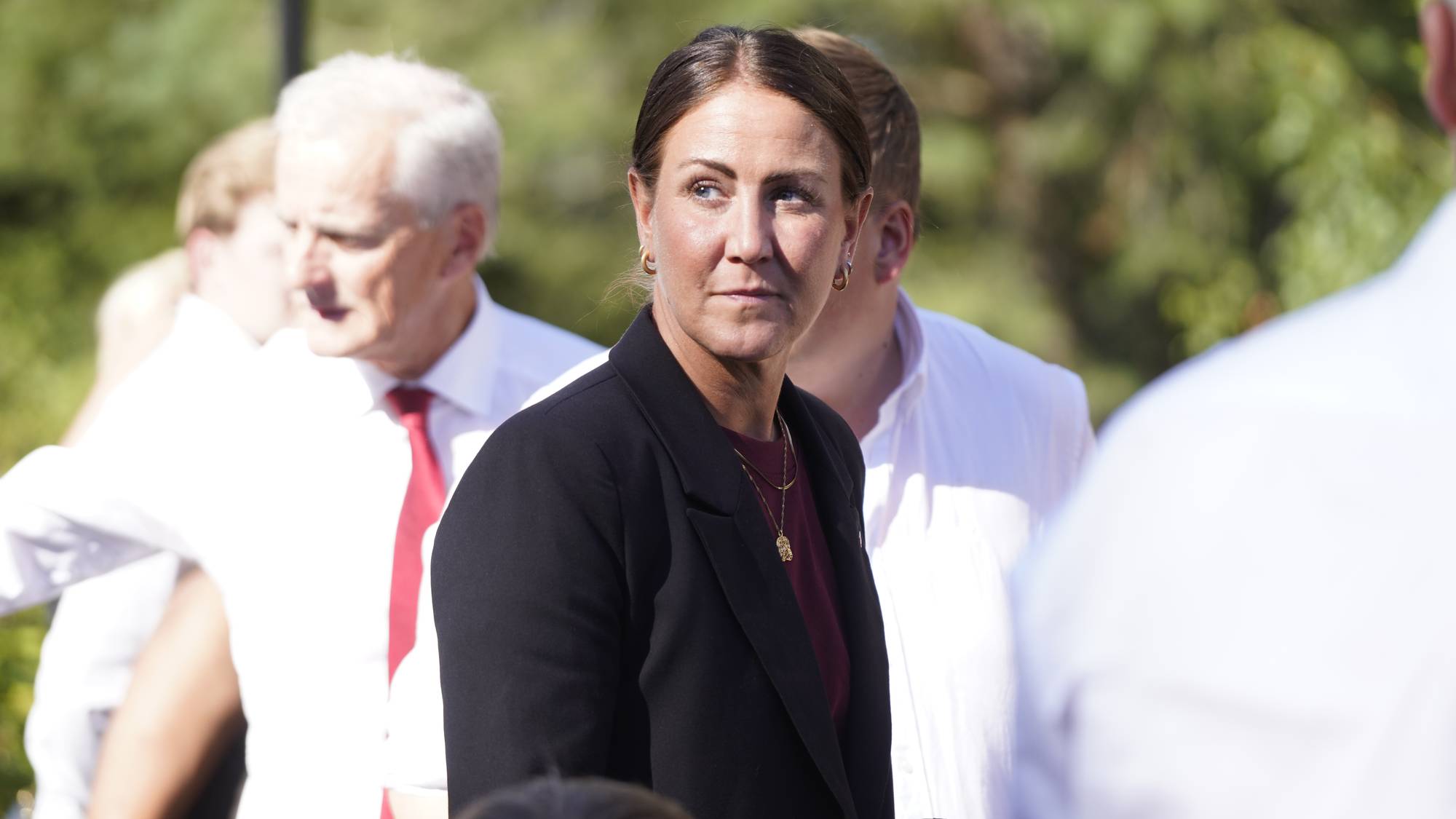 Kvinne i svart dressjakke står utendørs med Jonas Gahr Støre i bakgrunnen. (Bildebeskrivelsen er laget av en KI-tjeneste)