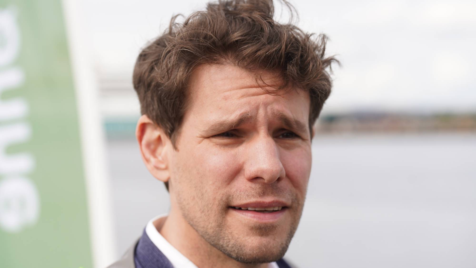 Et bilde av en mann i blå dressjakke. Mannen har brunt, rufsete hår og ser mot høyre i bildet. Han har en skjorte med knapp krage under jakken. I bakgrunnen er det et vann og en uskarp, grønn reklameplakat. (Bildebeskrivelsen er laget av en KI-tjeneste)