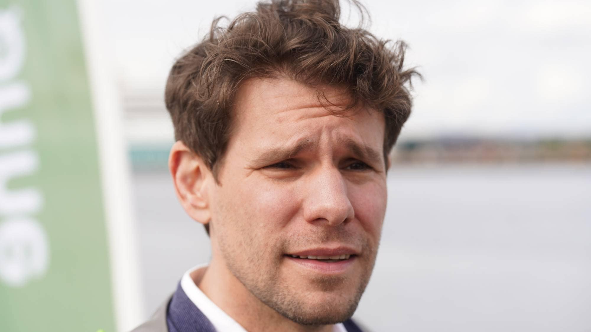 Et bilde av en mann i blå dressjakke. Mannen har brunt, rufsete hår og ser mot høyre i bildet. Han har en skjorte med knapp krage under jakken. I bakgrunnen er det et vann og en uskarp, grønn reklameplakat. (Bildebeskrivelsen er laget av en KI-tjeneste)