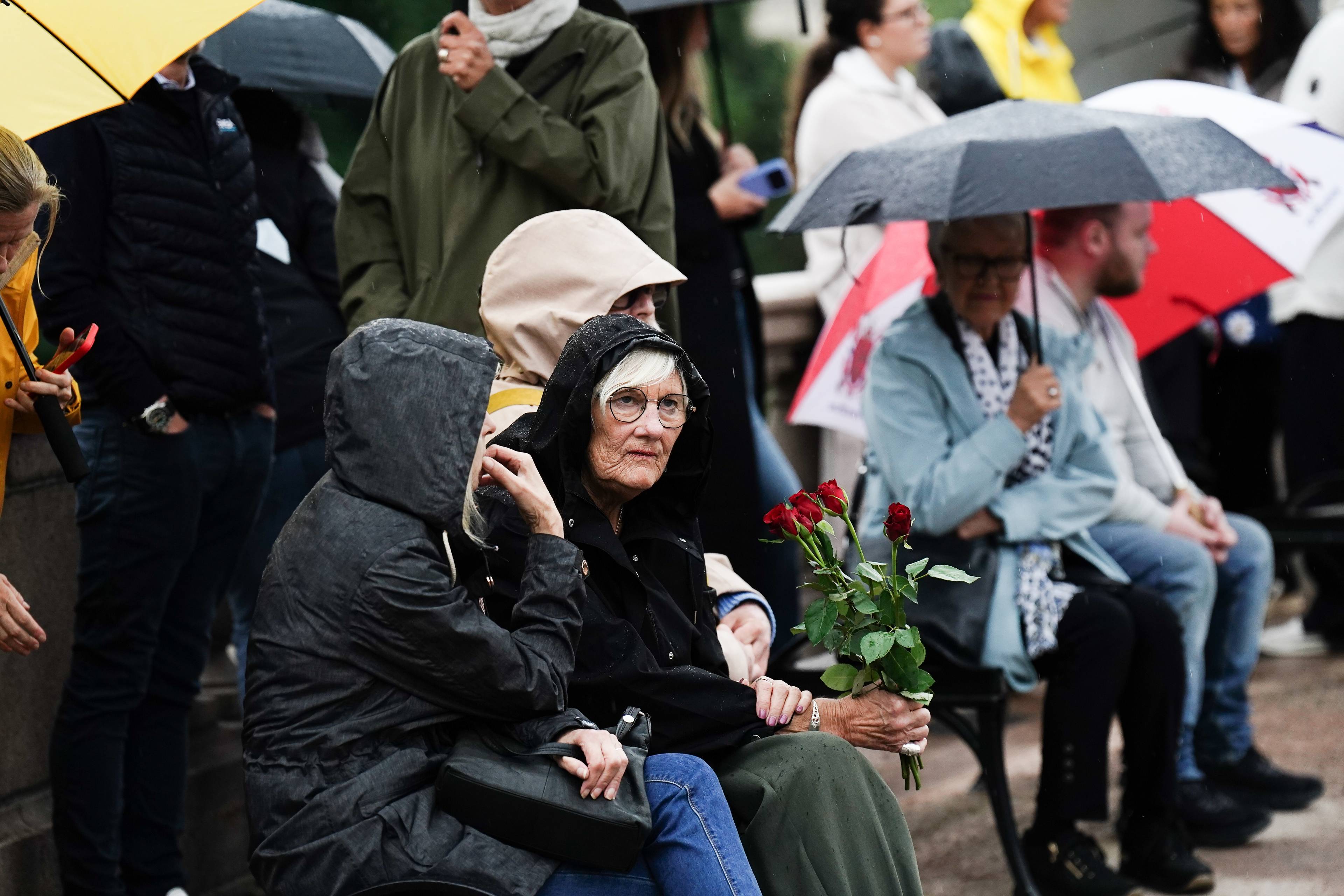 Folk med paraplyer sitter ute i regnet. En kvinne holder en bukett med røde roser. (Bildebeskrivelsen er laget av en KI-tjeneste)