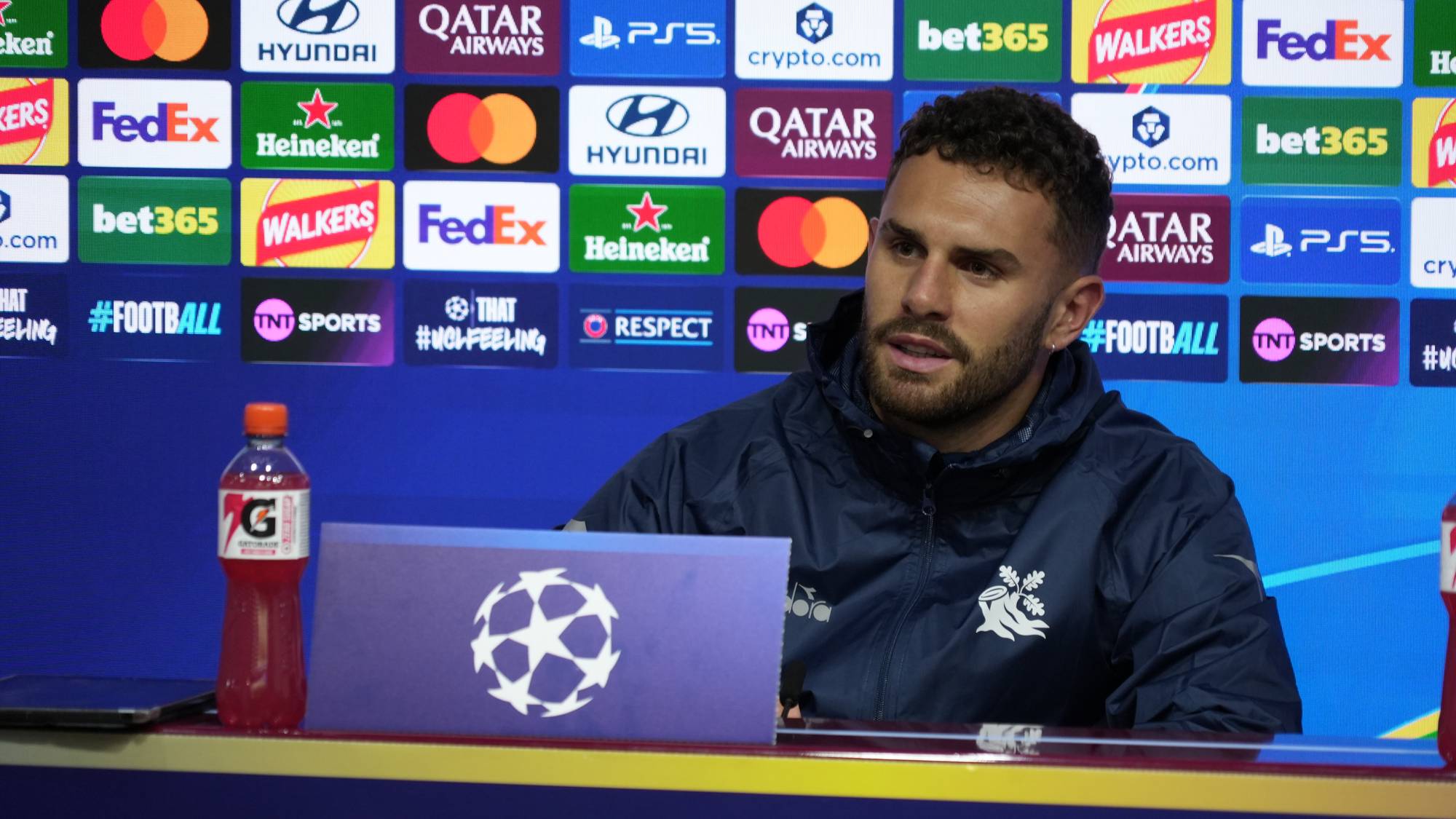 Fotballspiller sitter på pressekonferanse foran en reklamevegg med Champions League-logo. (Bildebeskrivelsen er laget av en KI-tjeneste)
