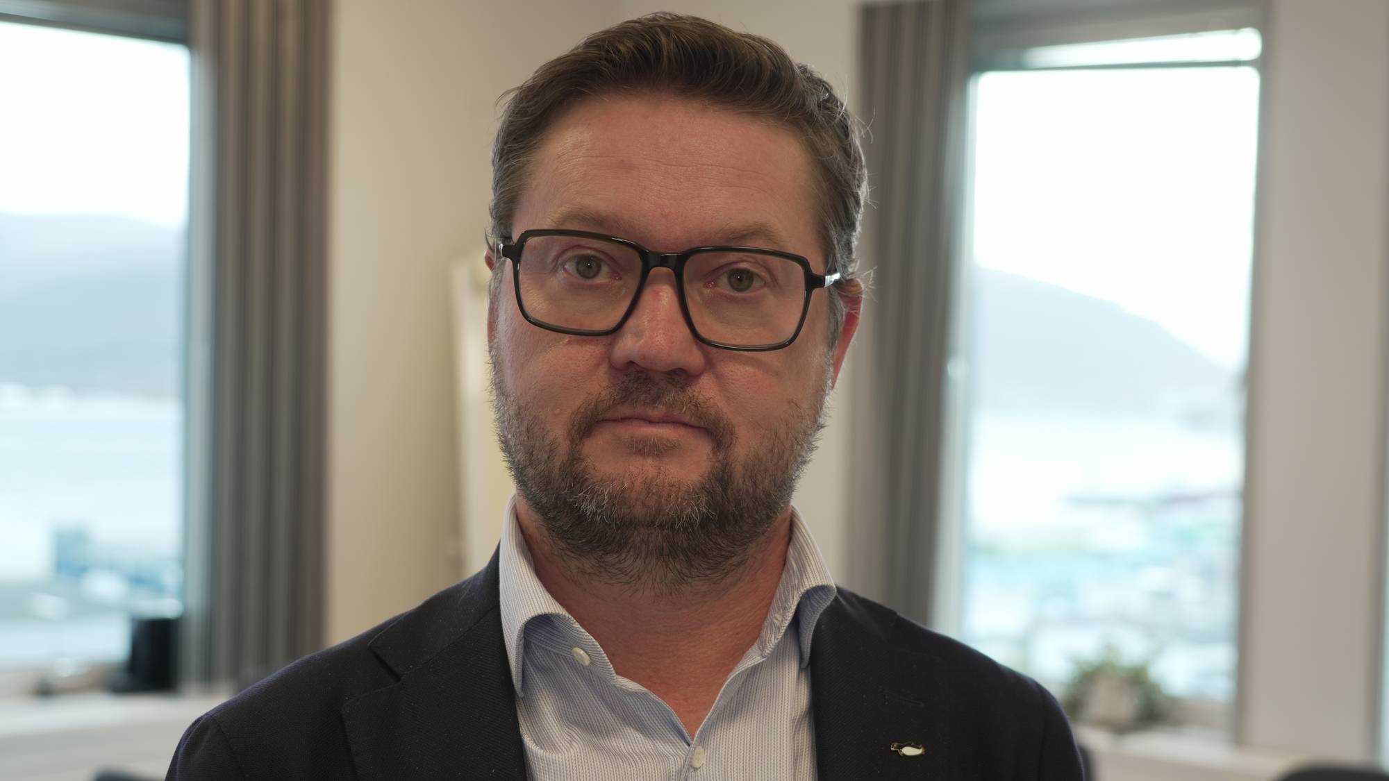 Portrett av en mann med briller og skjegg, kledd i skjorte og dressjakke, foran kontorvinduer. (Bildebeskrivelsen er laget av en KI-tjeneste)