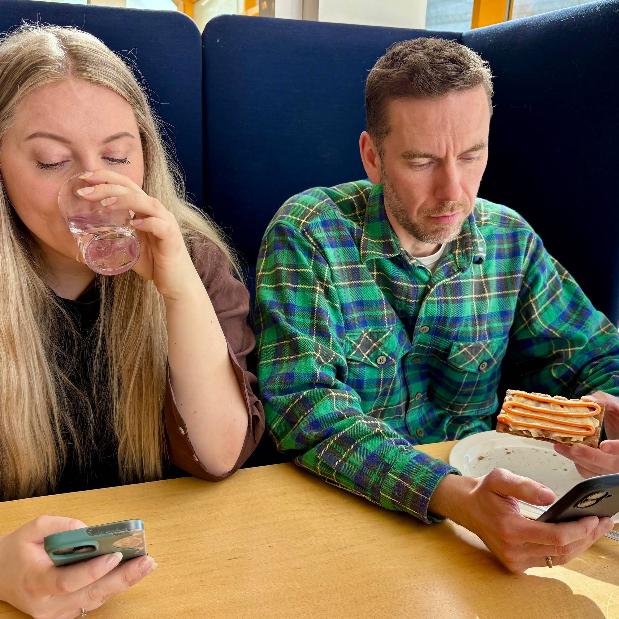 En dame og en mann sitter ved et bord og spiser lunsj. Begge ser på mobilen.
