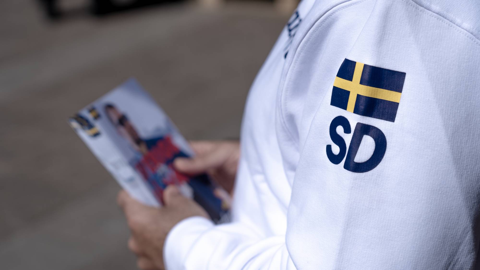 Person i hvit jakke med SD-logo og svensk flagg holder en brosjyre. (Bildebeskrivelsen er laget av en KI-tjeneste)