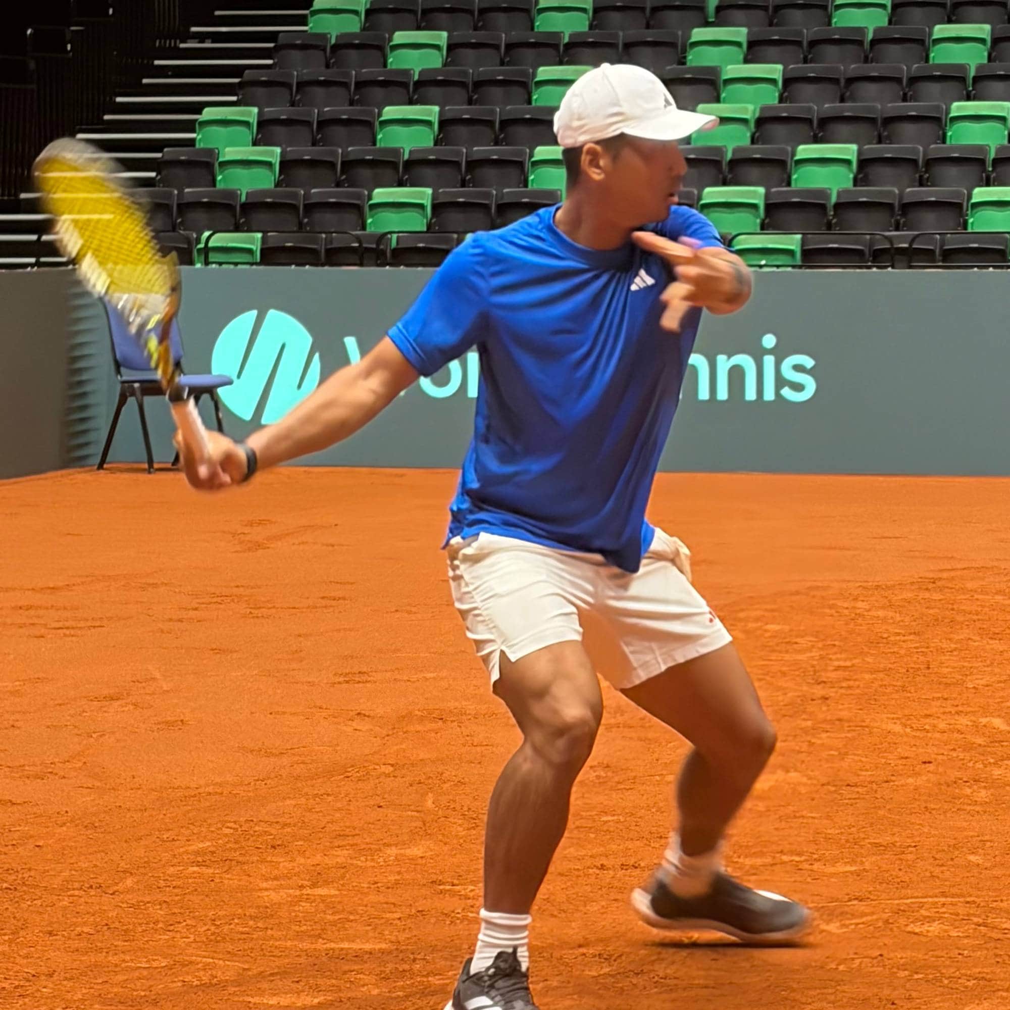 Tennisspiller i blå t-skjorte slår et slag på en grusbane i en tom hall. (Bildebeskrivelsen er laget av en KI-tjeneste)