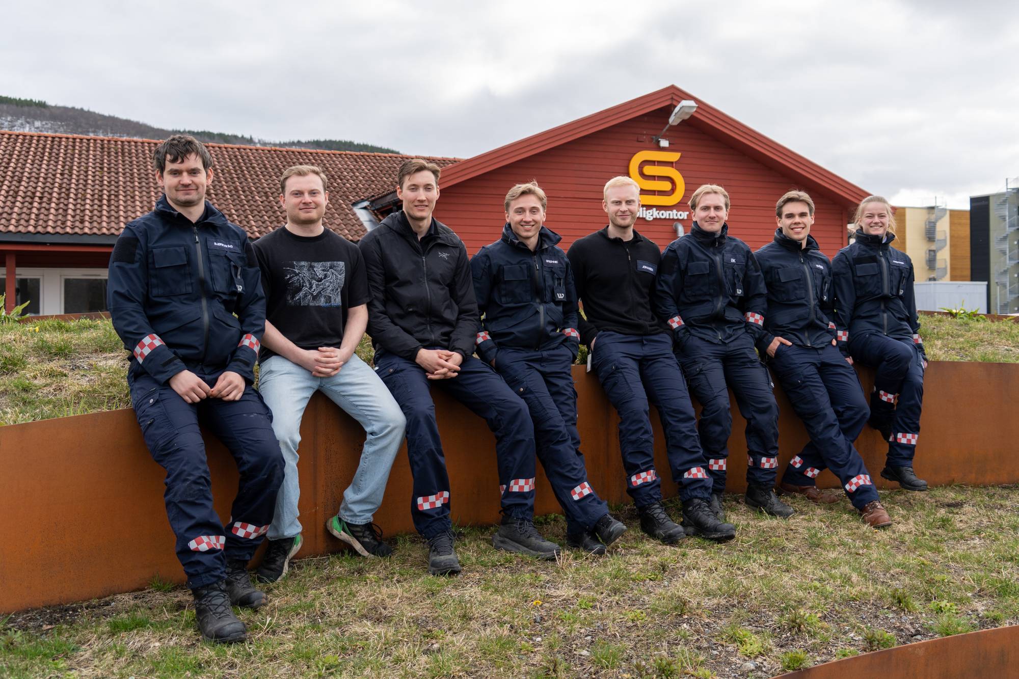 En gruppe på åtte personer sitter i en halvsirkel foran en rød bygning. Seks av personene er kledd i mørkeblå uniformer med reflekser. Bygningen har et stort gult S-symbol. De sitter på en lav vegg av cortenstål. (Bildebeskrivelsen er laget av en KI-tjeneste)