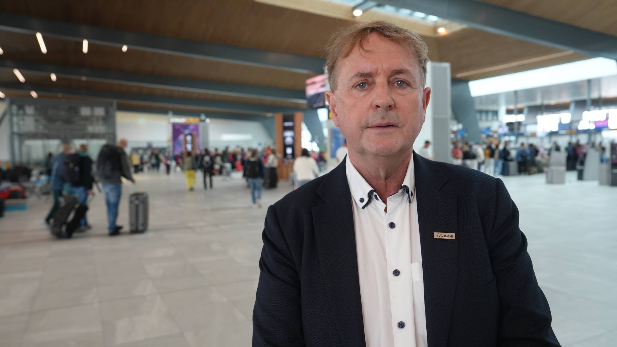 Mann i dress med Avinor-merke i en travel flyplasshall. (Bildebeskrivelsen er laget av en KI-tjeneste)