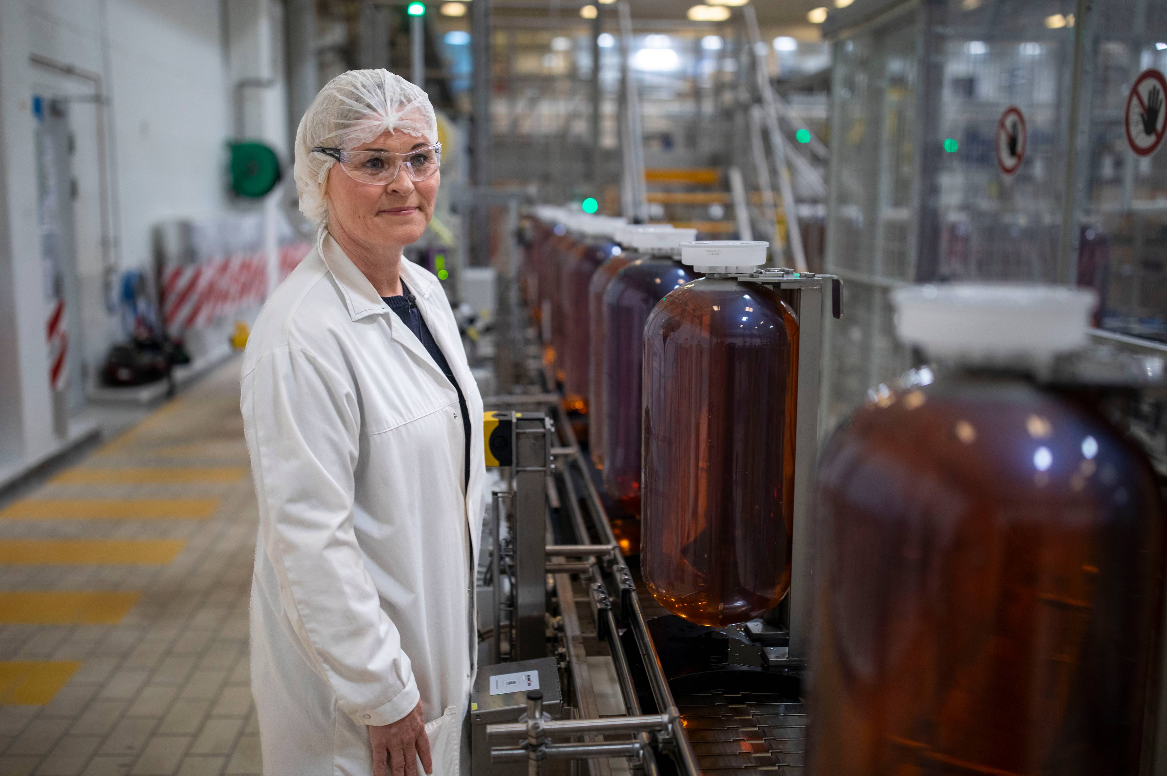 Kvinne i hvit frakk, briller og hårnett står på en fabrikk. Hun ser mot kameraet. Bak henne står store, brune beholdere på et transportbånd. Industrilokale med maskineri og lys.