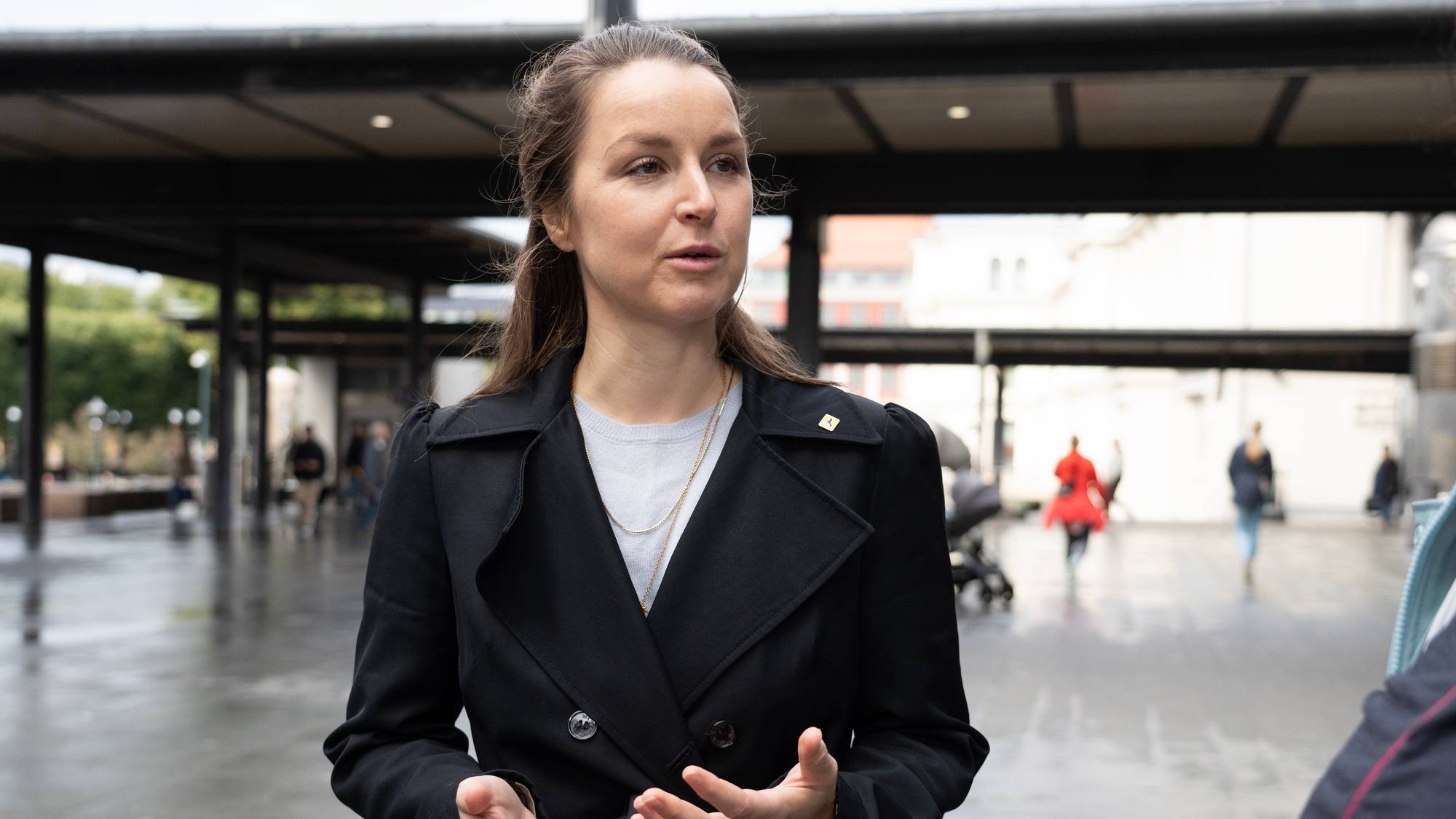 En kvinne står utendørs på et offentlig sted, og gestikulerer mens hun snakker. Hun har på seg en svart frakk og tilbehør som en kjede. I bakgrunnen kan man se mennesker gå forbi, noen med barnevogn, og bygninger som gir en urban kontekst. Det er vått på bakken, noe som tyder på nylig nedbør. (Bildebeskrivelsen er laget av en KI-tjeneste)