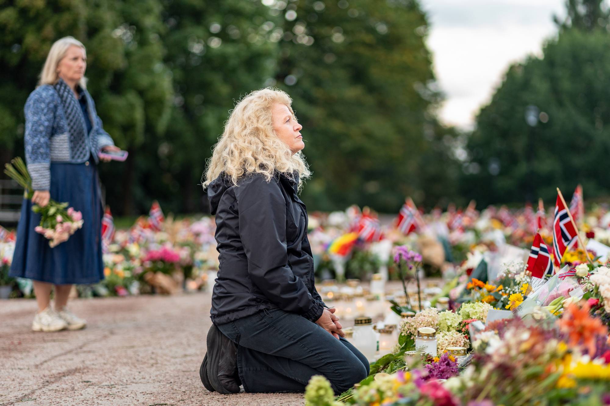 Kvinne kneler foran et blomsterhav med norske flagg. En annen kvinne står bak med blomster. (Bildebeskrivelsen er laget av en KI-tjeneste)