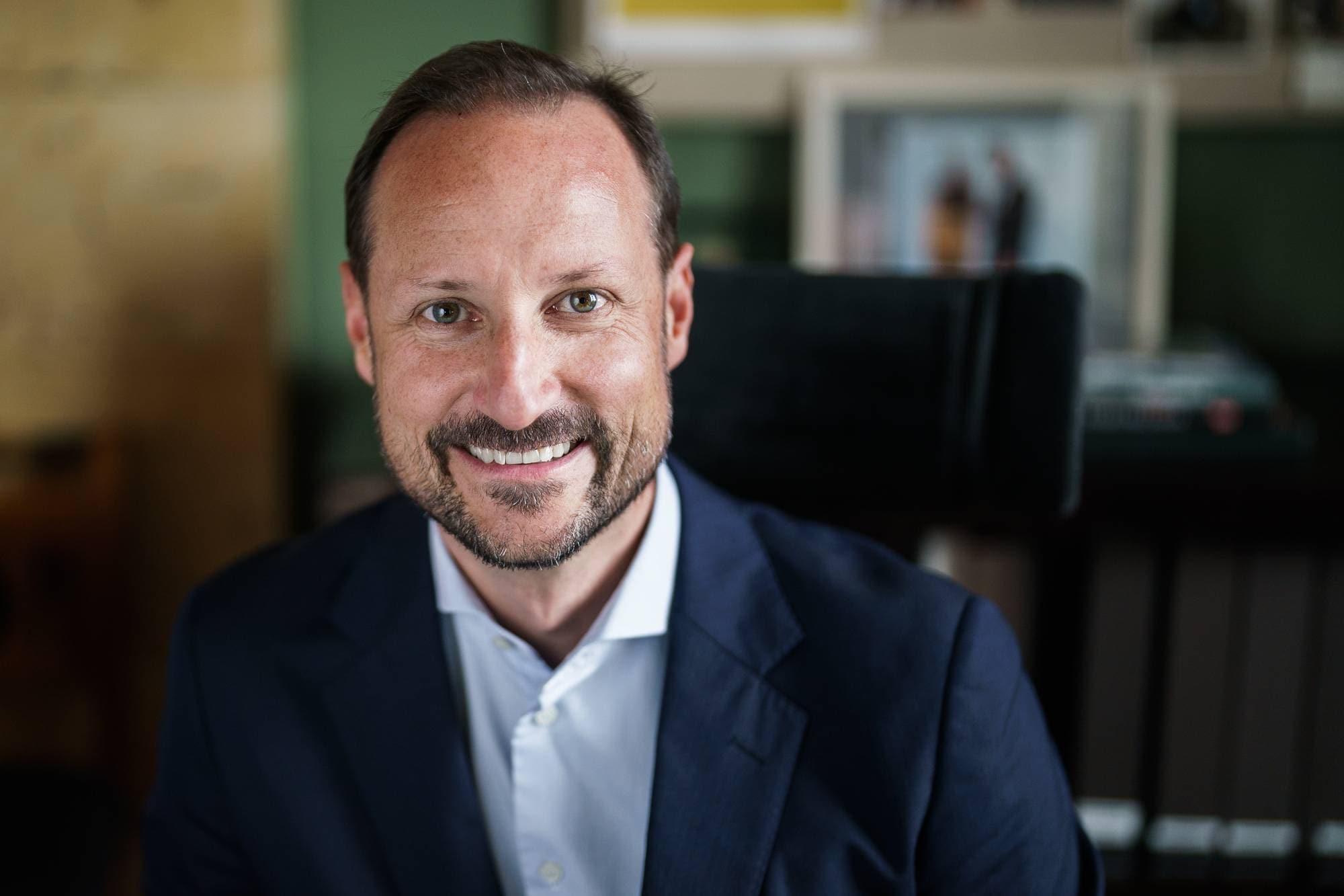 Kronprins Haakon ser inn i kamera og smiler mens han sitter på kontoret sitt