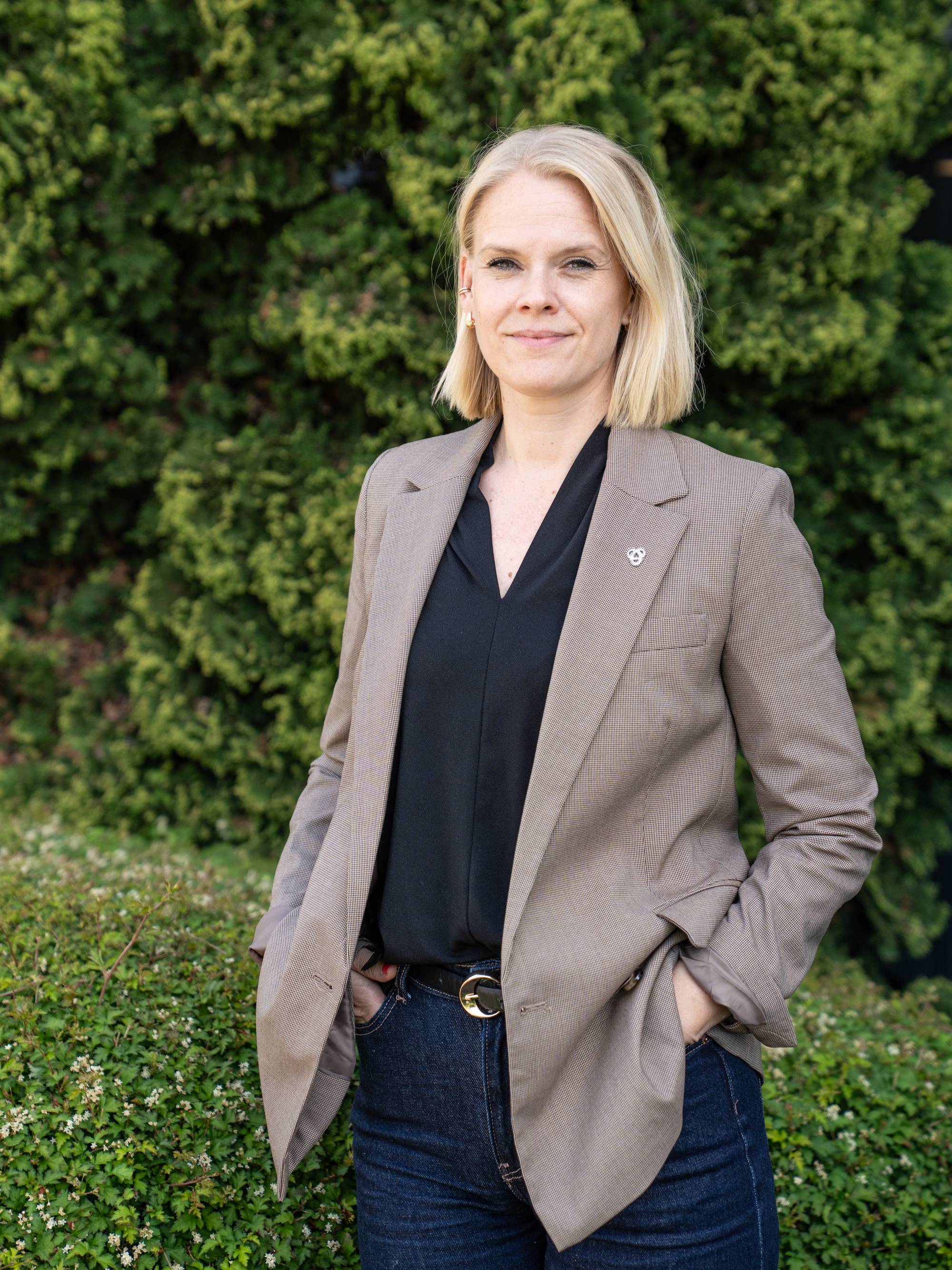 En kvinne med blondt hår står foran en hekk og et grønt tre. Hun har på seg en blazer og mørke jeans. Kvinnen har hendene i lommene. (Bildebeskrivelsen er laget av en KI-tjeneste)