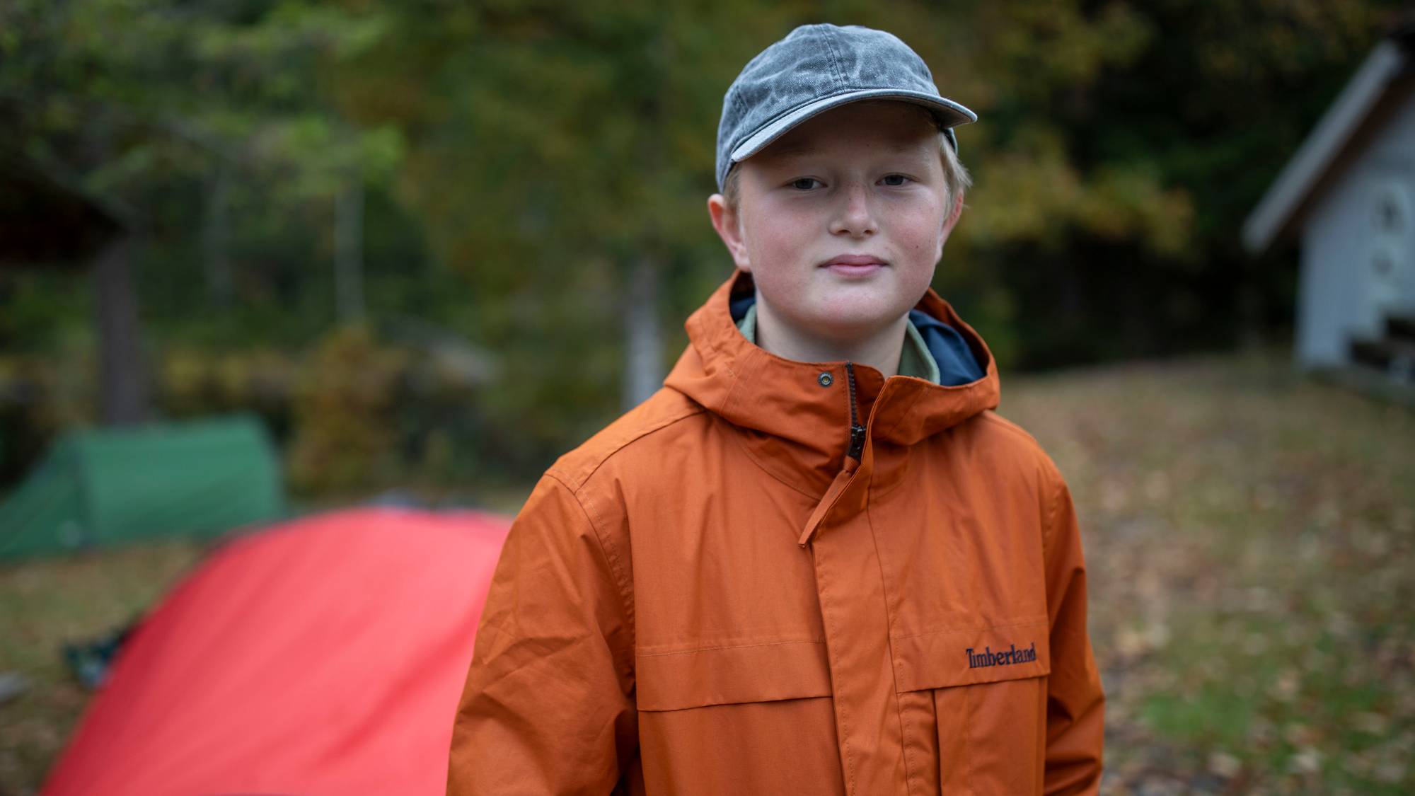En gutt med caps og oransje jakke står ute ved telt i skogen. (Bildebeskrivelsen er laget av en KI-tjeneste)