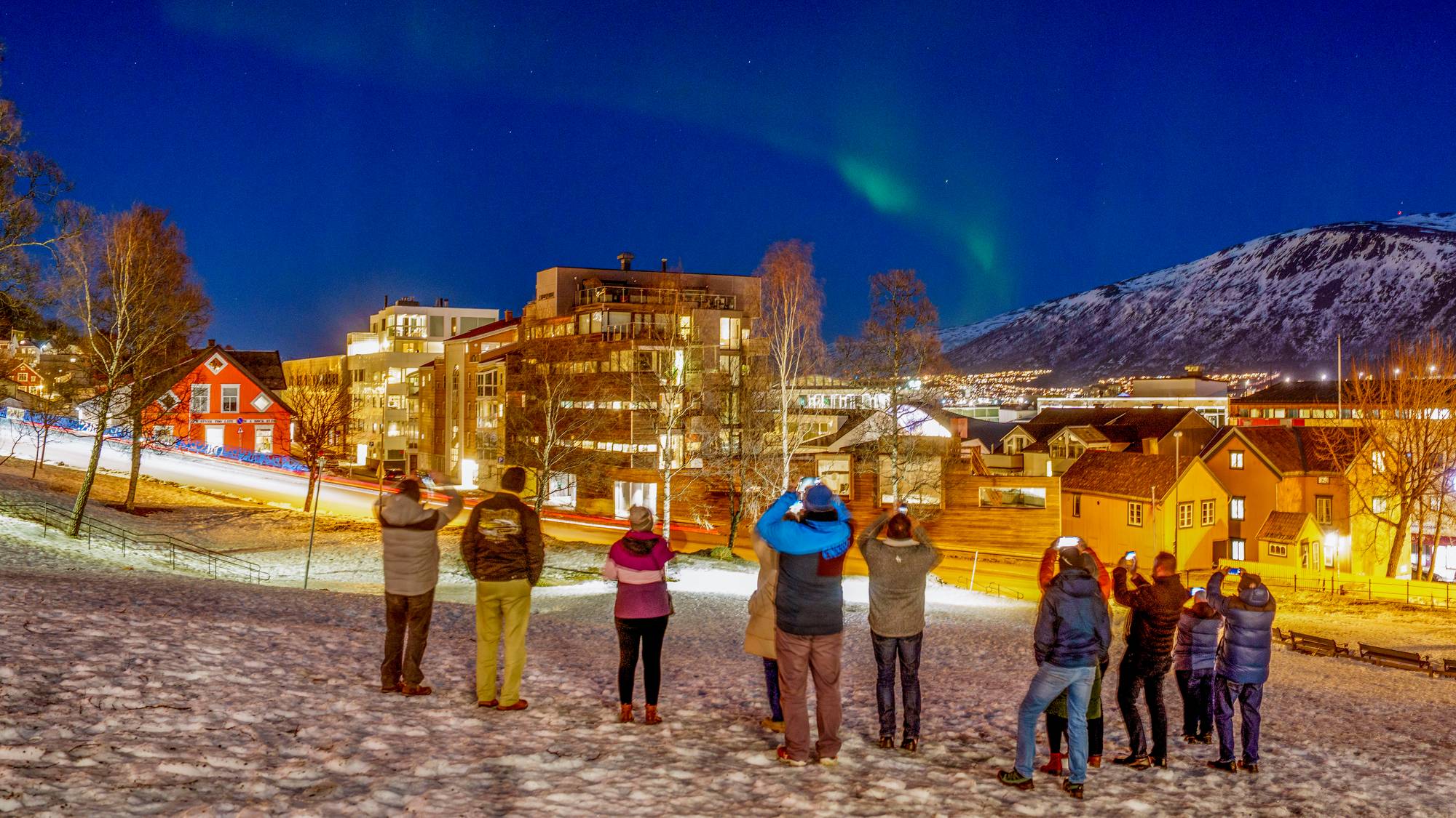 En gruppe turister tar bilder av nordlys, de står på en snødekt bakke med bygninger og fjell i bakgrunnen.