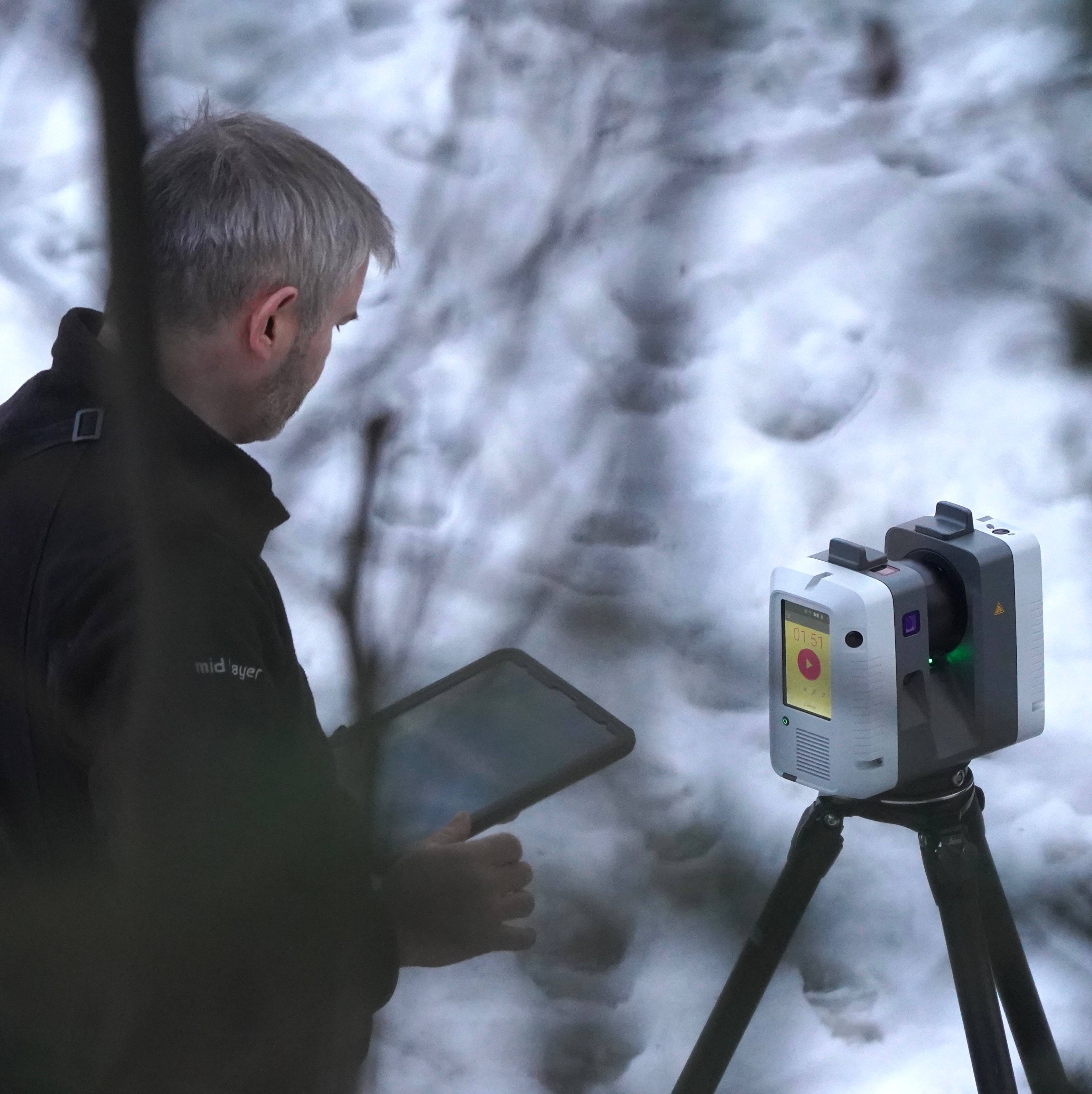 En mann med nettbrett ser på en 3D-skanner. Skanneren er montert på et stativ. Begge befinner seg ute i snødekt natur. Bakgrunnen er uskarp og viser trær og snø.