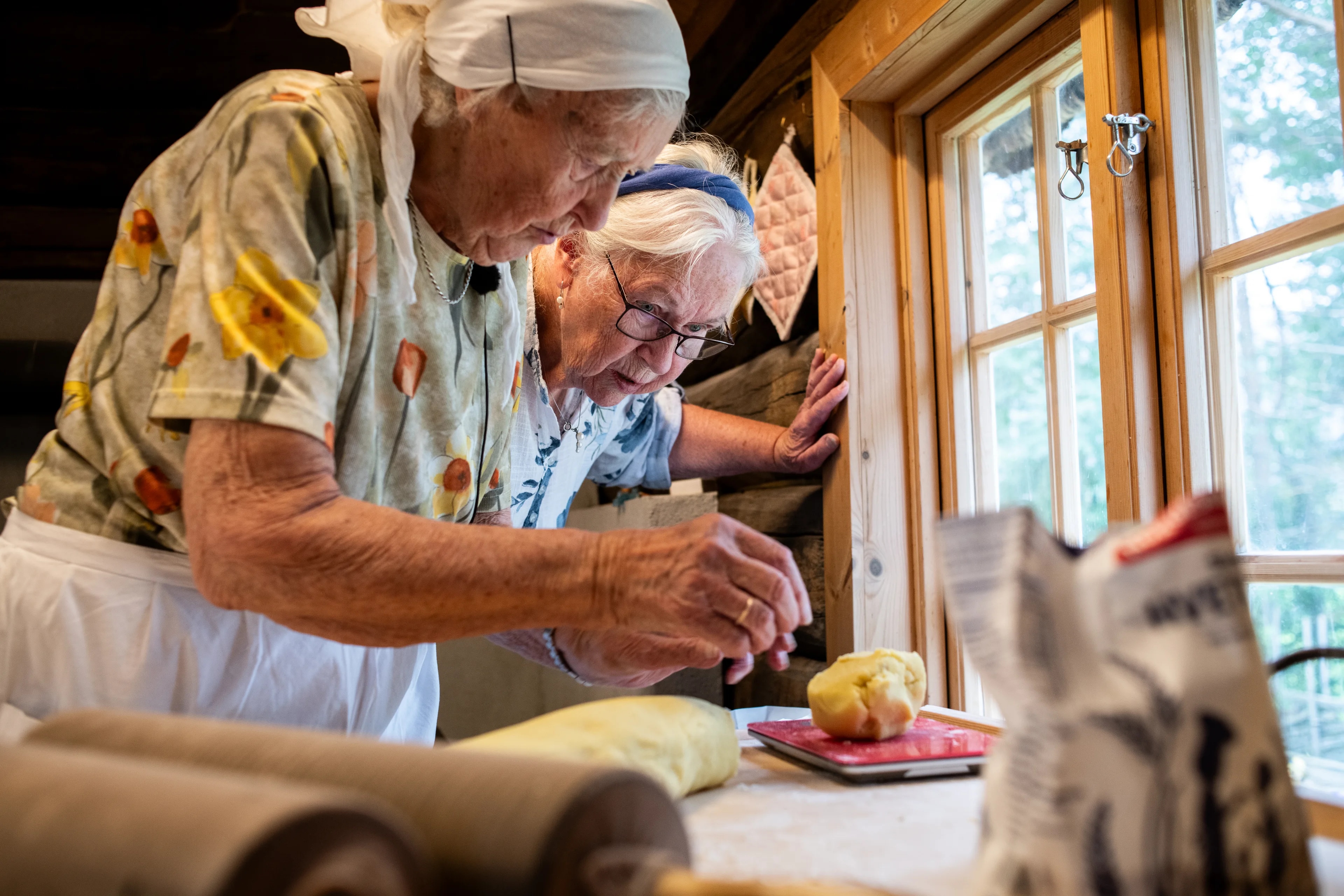 To eldre kvinner baker sammen ved et vindu i et tømmerhus.