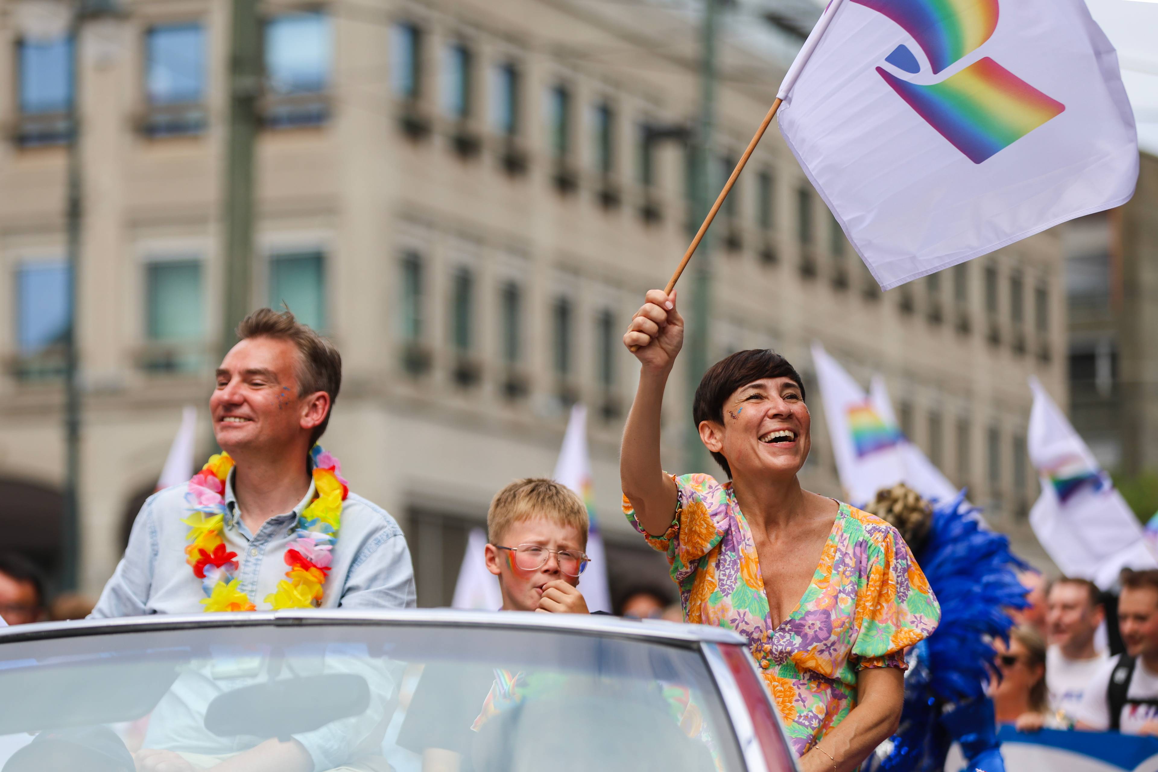 Kvinne vifter med regnbueflagg fra en åpen bil under Pride-parade sammen med mann og barn. (Bildebeskrivelsen er laget av en KI-tjeneste)