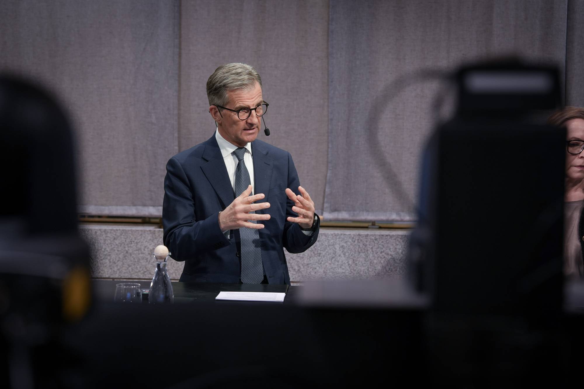 En mann i dress holder en presentasjon.
Han har briller og en mikrofon festet til jakkeslaget.
Mannen står bak et bord med en vannflaske og et glass.
Bak ham er det en grå bakgrunn.