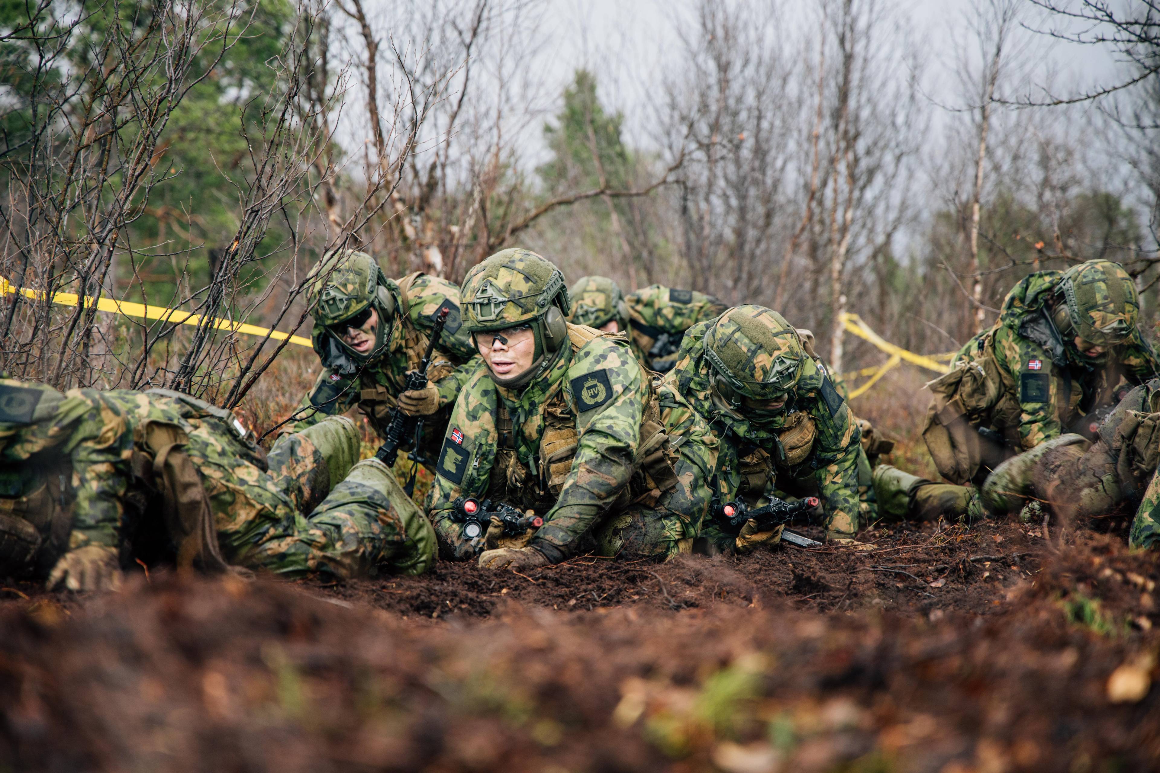 Fem kvinnelige soldater kryper gjennom gjørme under øvelse.  Det er høst. I midten soldat med hjelp og briller. På ermet sees HV-merke.