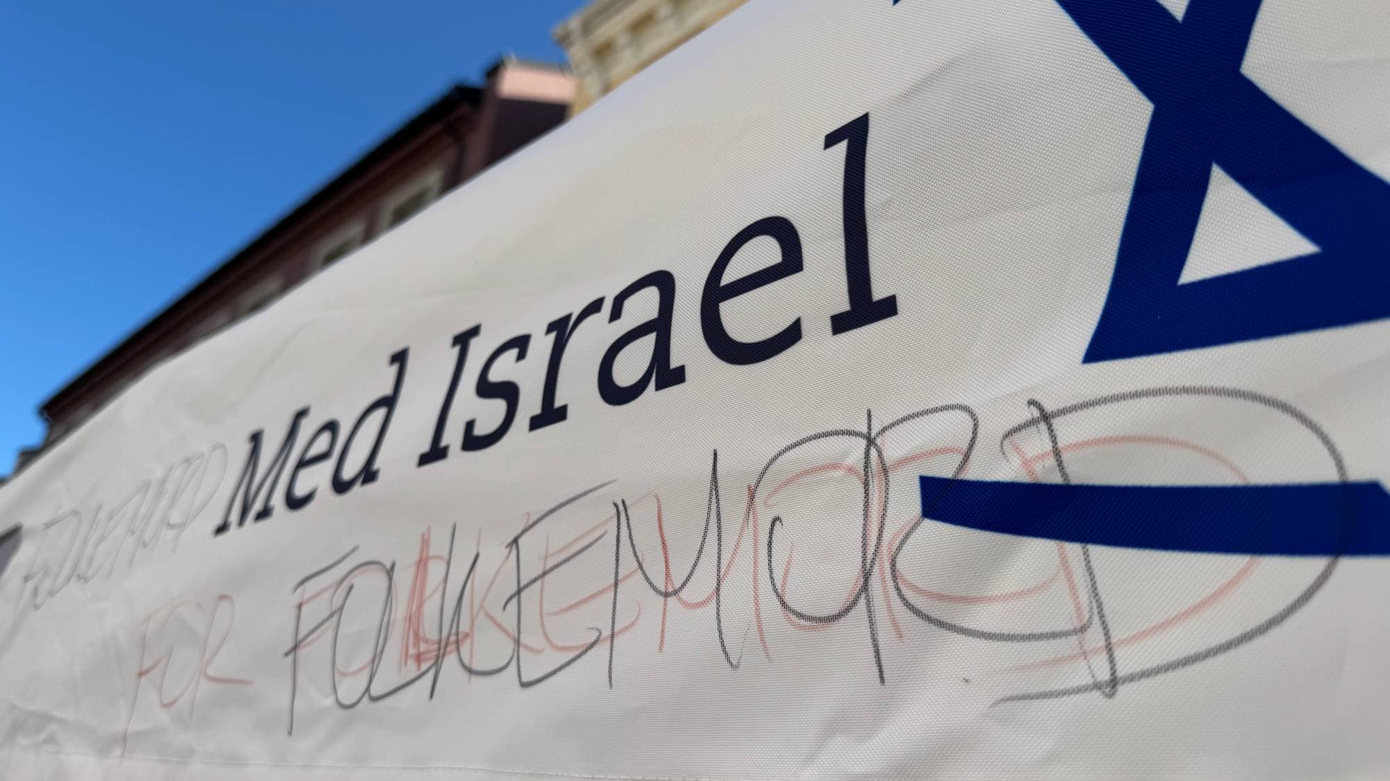Banner med teksten Med Israel og påført tekst om folkemord, sett nedenfra mot en blå himmel. (Bildebeskrivelsen er laget av en KI-tjeneste)