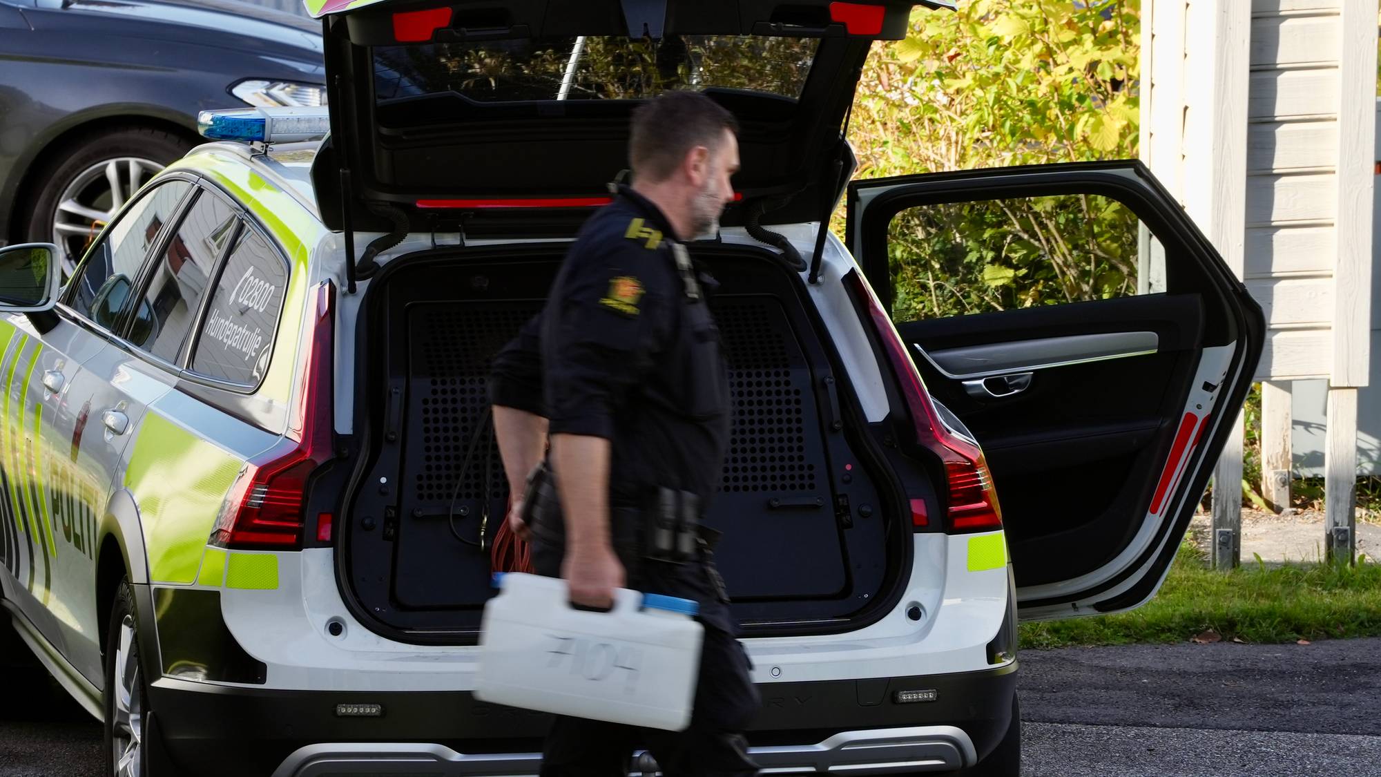 En politibetjent går fra en politibil med åpent bagasjerom og bærer en hvit plastbeholder. (Bildebeskrivelsen er laget av en KI-tjeneste)
