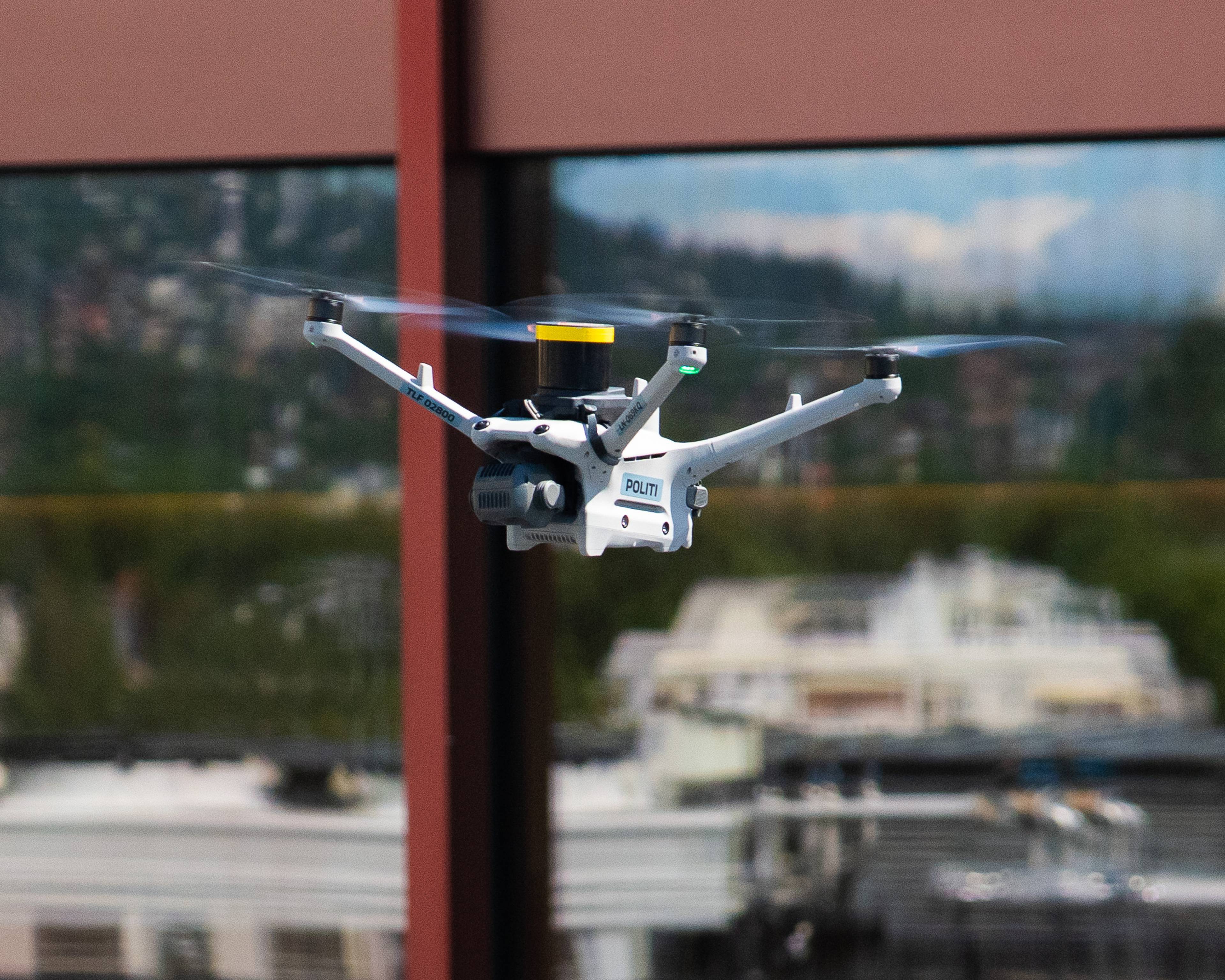 En politidrone flyr foran en bygning. Dronen er hvit med et gult element på toppen og har inskripsjonen "POLITI". I bakgrunnen er det et speilende vindu som reflekterer en skyfull himmel og trær. Den røde søylen til venstre og høyre antyder et moderne arkitektonisk byggverk. (Bildebeskrivelsen er laget av en KI-tjeneste)