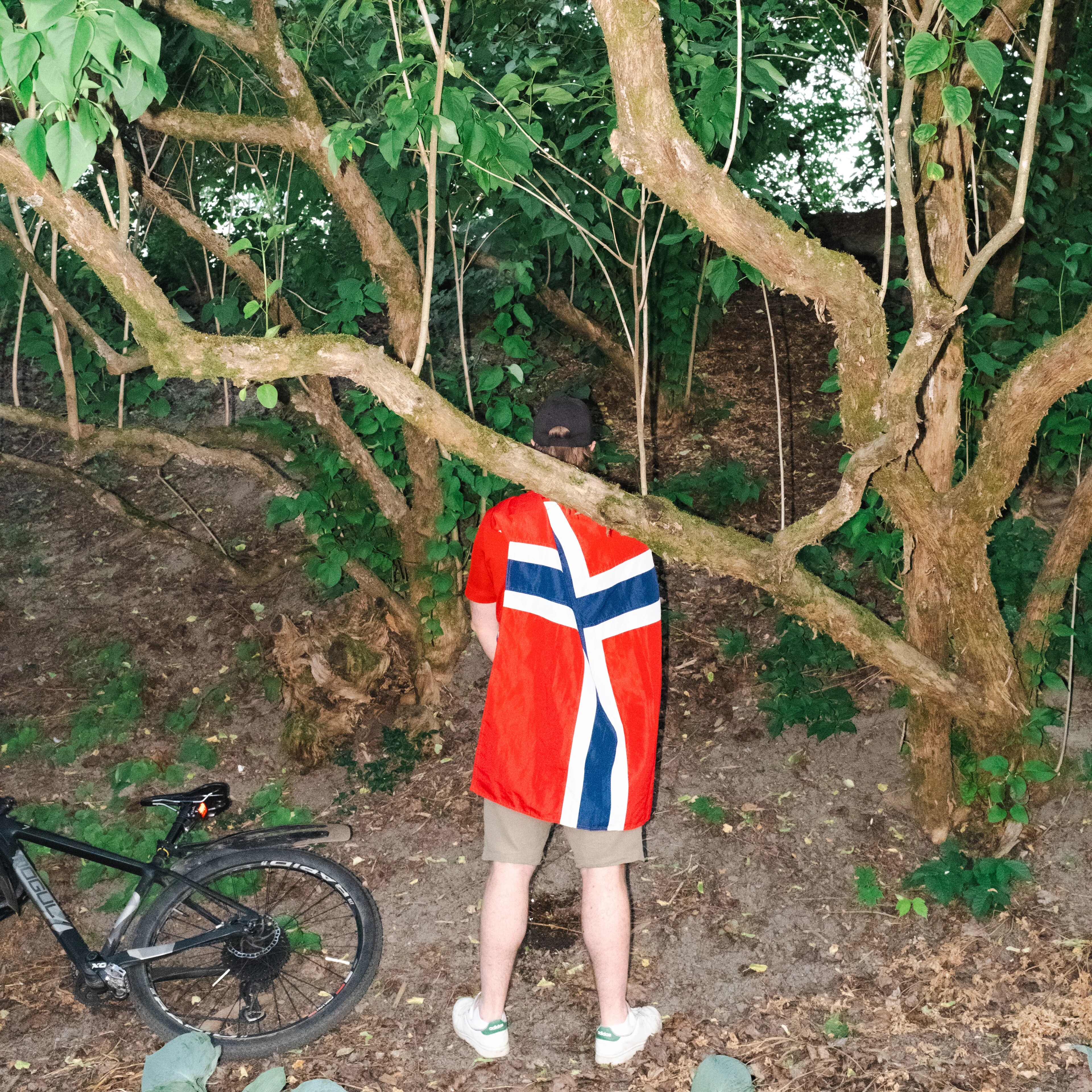 Person med et norsk flagg som kappe står foran et tre i skogen med en sykkel ved siden av seg.