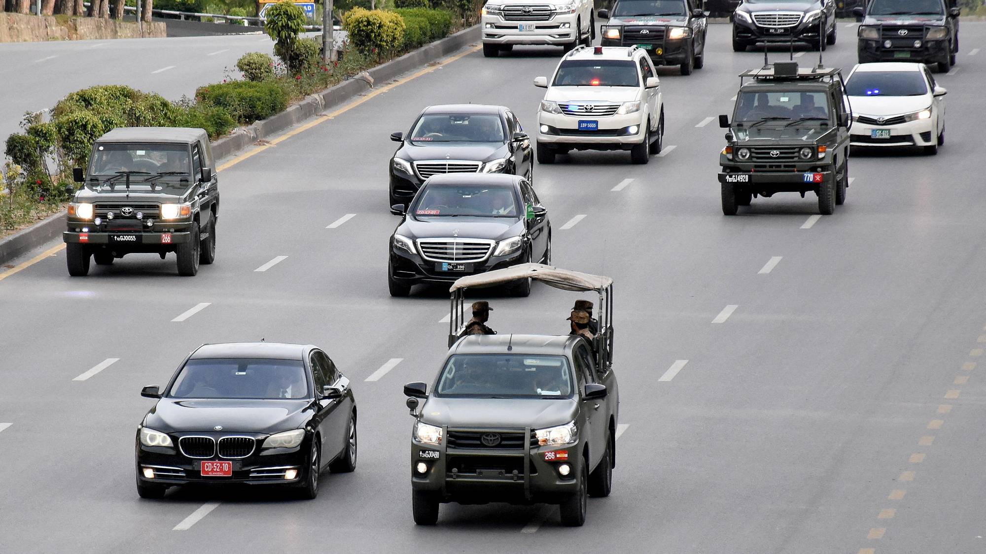En bilkolonne med flere kjøretøy, inkludert SUV-er og sedaner, kjører på en vei. Blant bilene er det en åpen militær-jeep med to personer i. Veien er omgitt av trær og grøntområder. (Bildebeskrivelsen er laget av en KI-tjeneste)