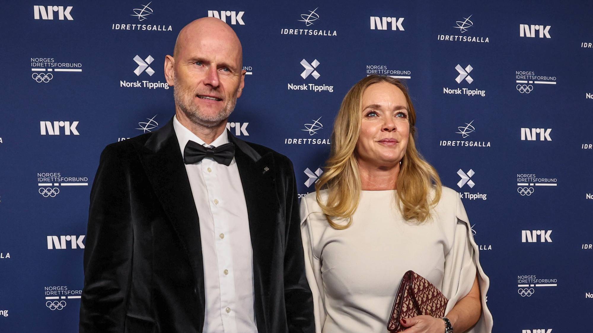 To personer står hånd i hånd på den røde løperen, iført festklær. Mannen har på seg en svart dress med flosshatt, mens kvinnen er iført en lys kjole. Bakgrunnen viser et banner med ulike sponsorer og arrangementets navn. Bildet fanger en elegant atmosfære typisk for et offentlig arrangement. (Bildebeskrivelsen er laget av en KI-tjeneste)