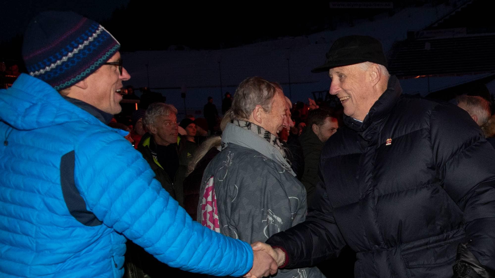 Kong Harald håndhilser på en person i blå jakke utendørs på kveldstid. (Bildebeskrivelsen er laget av en KI-tjeneste)