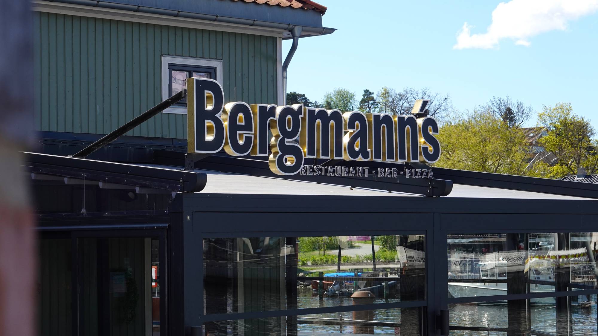 En bygning med skiltet "Bergmanns Restaurant - Bar - Pizza". Det er en sommerdag med blå himmel og skyer. Bygningen ligger ved vann, og det er båter i vannet. (Bildebeskrivelsen er laget av en KI-tjeneste)