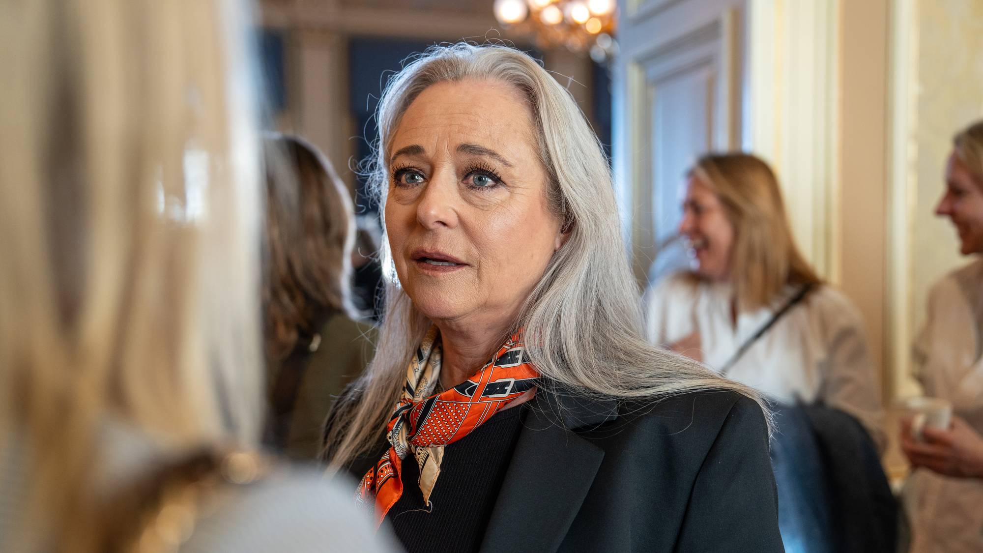 En kvinne med grått hår drikker vann og smiler. Rundt henne står mange kvinner. 
