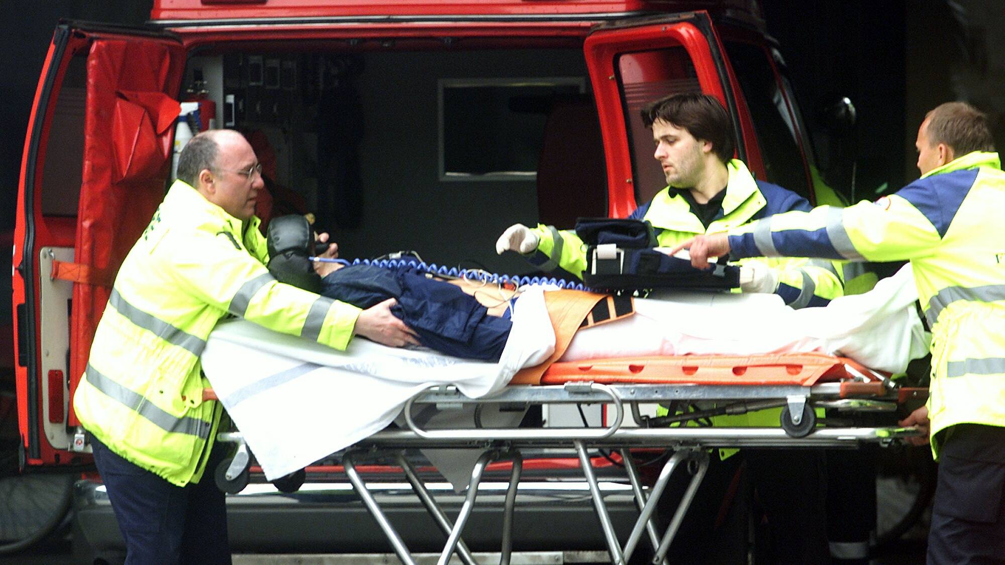 Ambulansepersonell legger en pasient på båre inn i ambulanse. Tre personer i gule varseljakker er synlige. Bakgrunnen er bakdørene på en rød ambulanse. (Bildebeskrivelsen er laget av en KI-tjeneste)