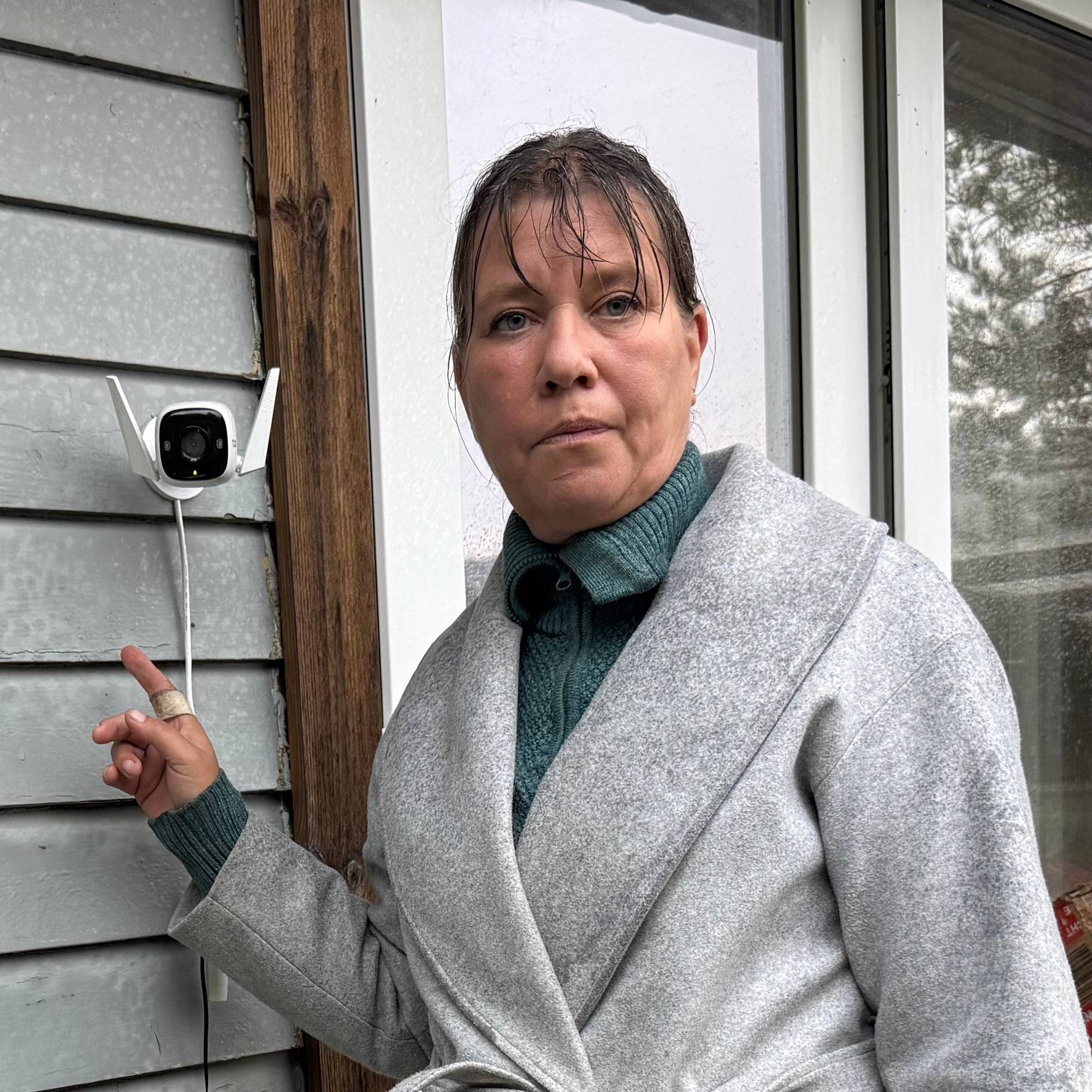 Nina Olsen Holm, kvinne med grå genser, står ved inngangsdøra, viser frem overvåkningskamera