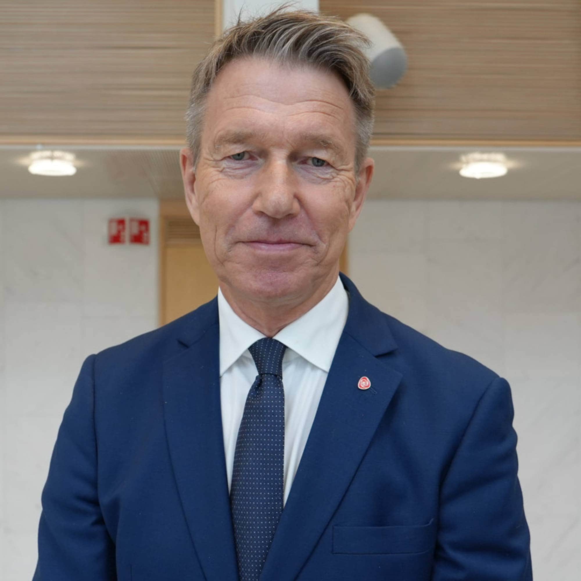 Portrett av en mann i blå dress og slips stående foran en lys vegg i en offentlig bygning. (Bildebeskrivelsen er laget av en KI-tjeneste)