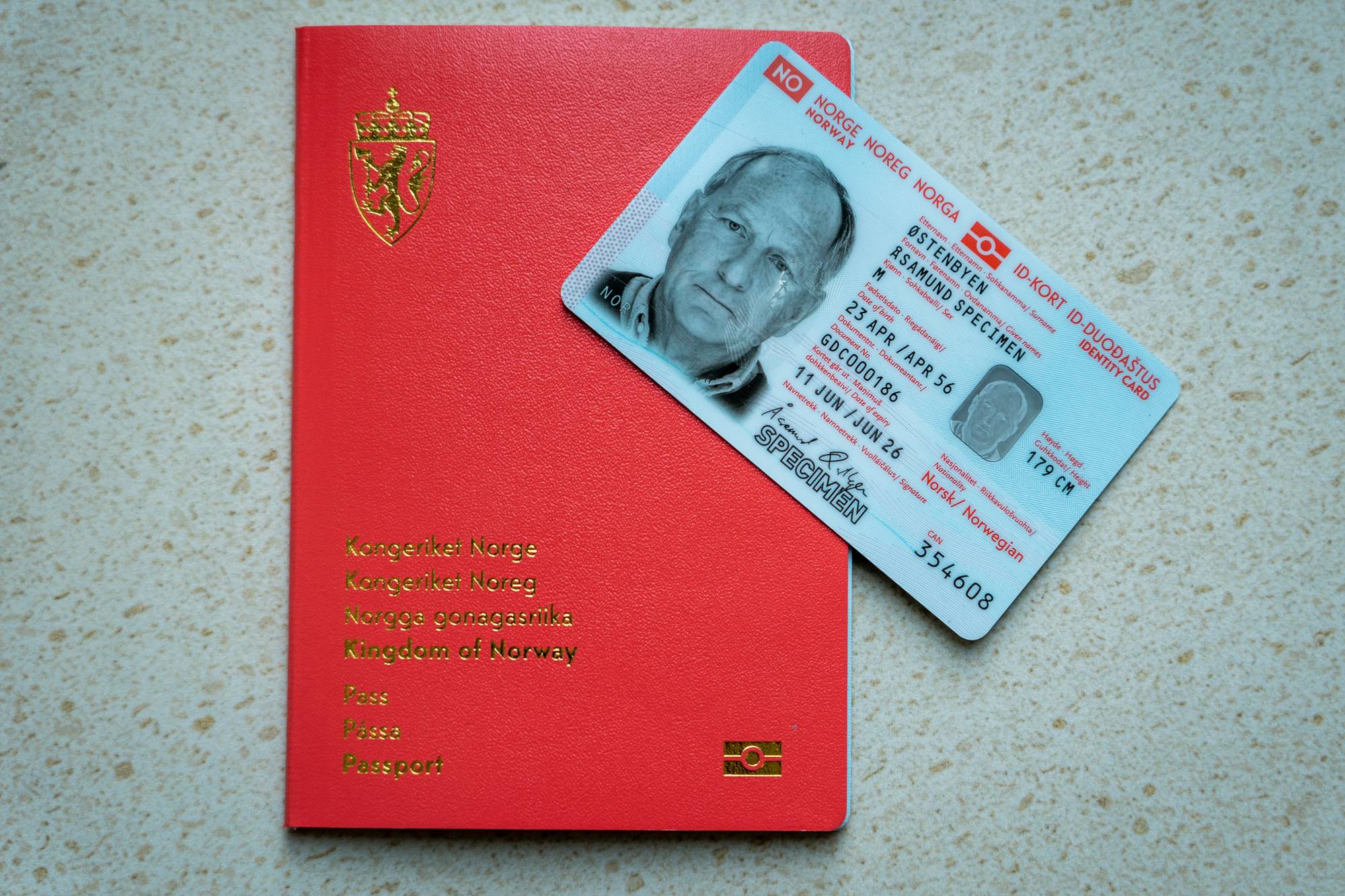 Et rødt norsk pass og et norsk ID-kort ligger ved siden av hverandre. ID-kortet har et bilde av en eldre mann og teksten "SPECIMEN" over ansiktet hans. Passet har et gullfarget riksvåpen og teksten "Kongeriket Norge" på forsiden. (Bildebeskrivelsen er laget av en KI-tjeneste)