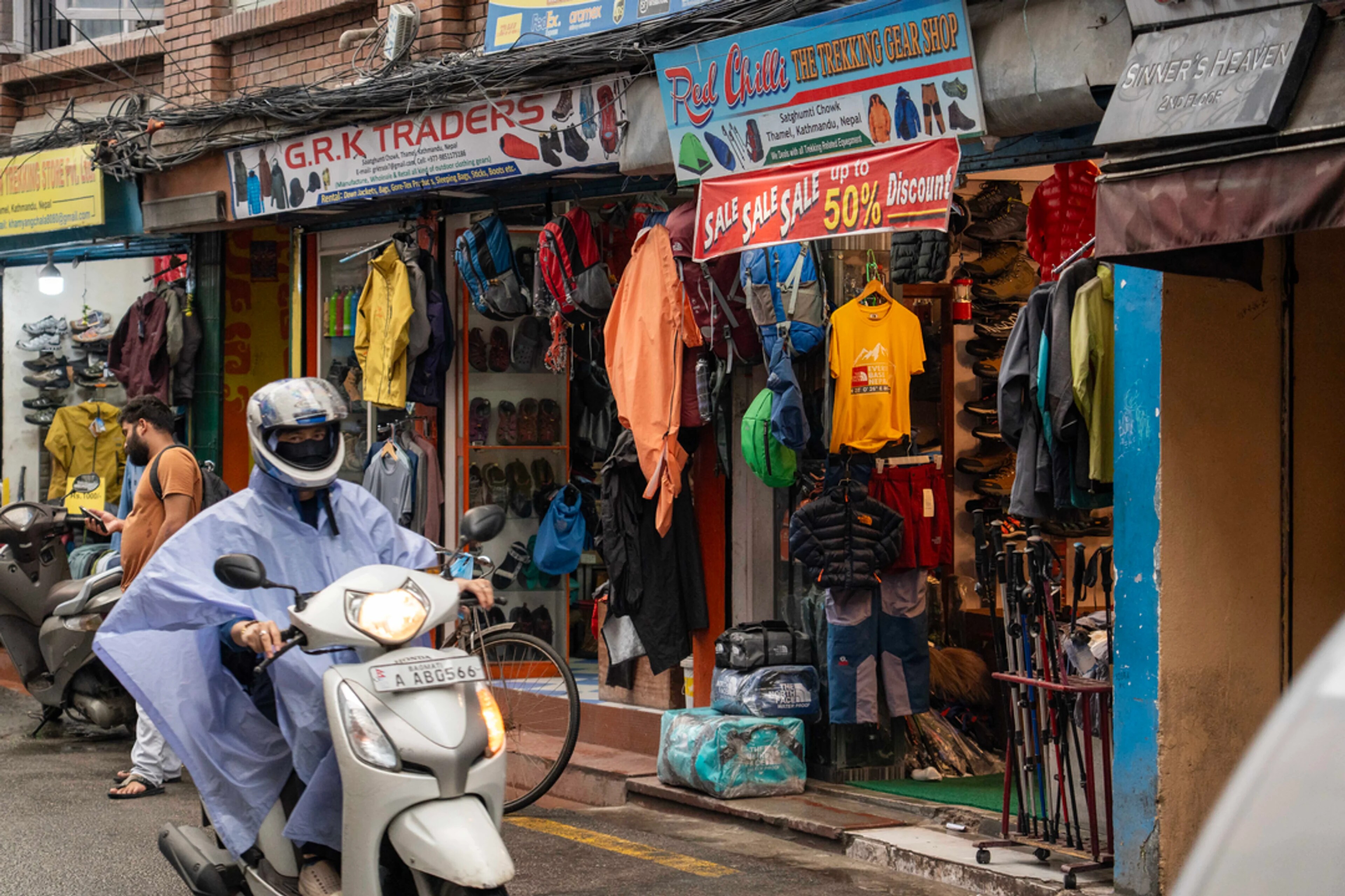 Mann på moped i Katmandu – Person i lyseblå regndress på kvit moped, kvit hjelm. Butikk med jakker og T-skjorter som henger på fasaden.