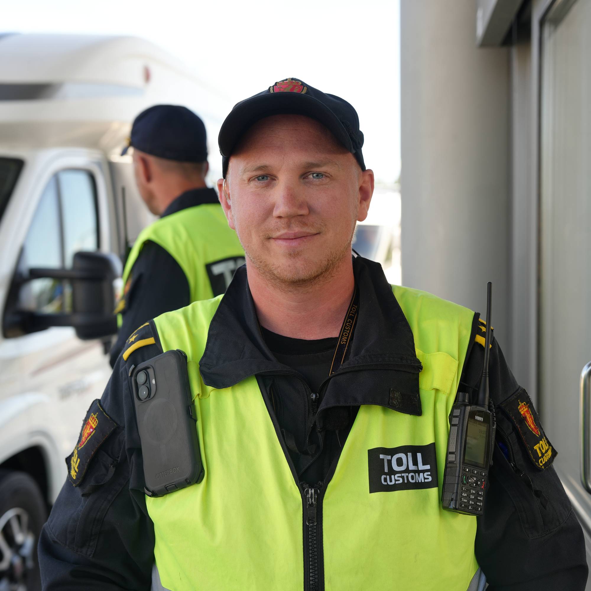 Tollbetjent i uniform står foran et kjøretøy på en tollstasjon. (Bildebeskrivelsen er laget av en KI-tjeneste)