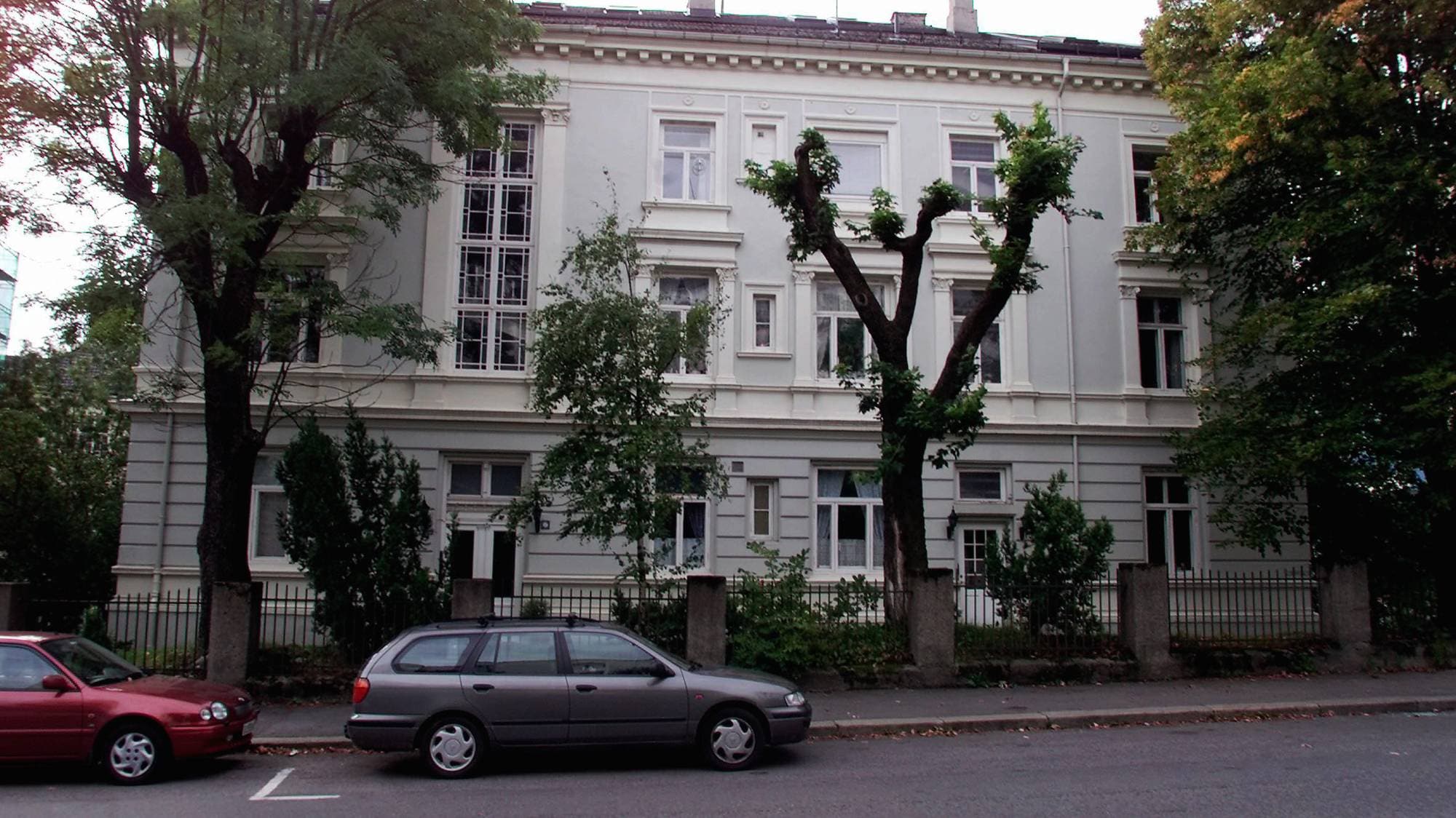 Fasaden på en grå bygård. I veien er to biler parkert, og det er trær rundt bygården.