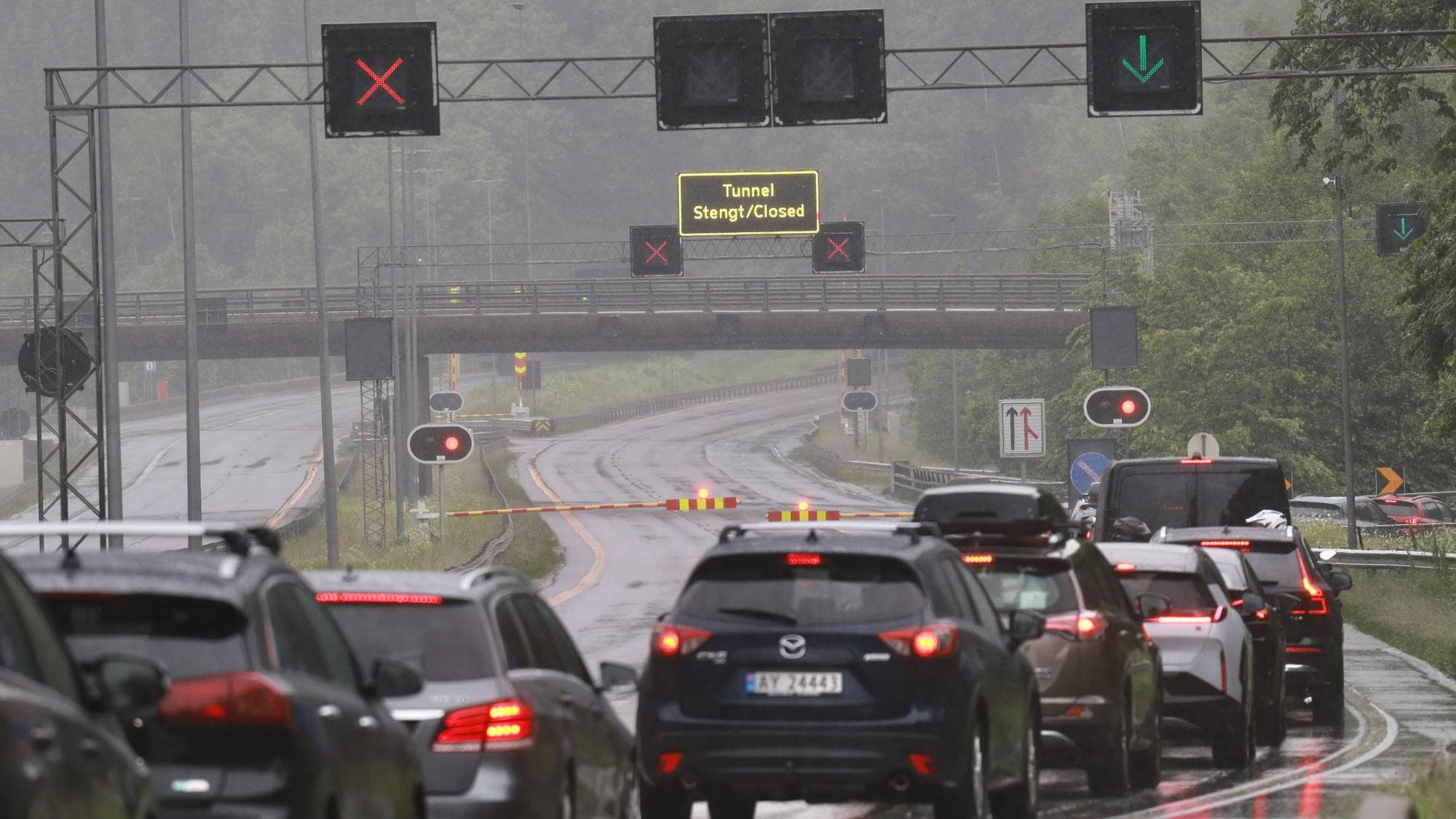 Biler står i kø på en våt vei foran et tunnel som er stengt. Trafikklys viser rødt, og en bom er nede. Et digitalt skilt over veien viser teksten "Tunnel Stengt/Closed". 