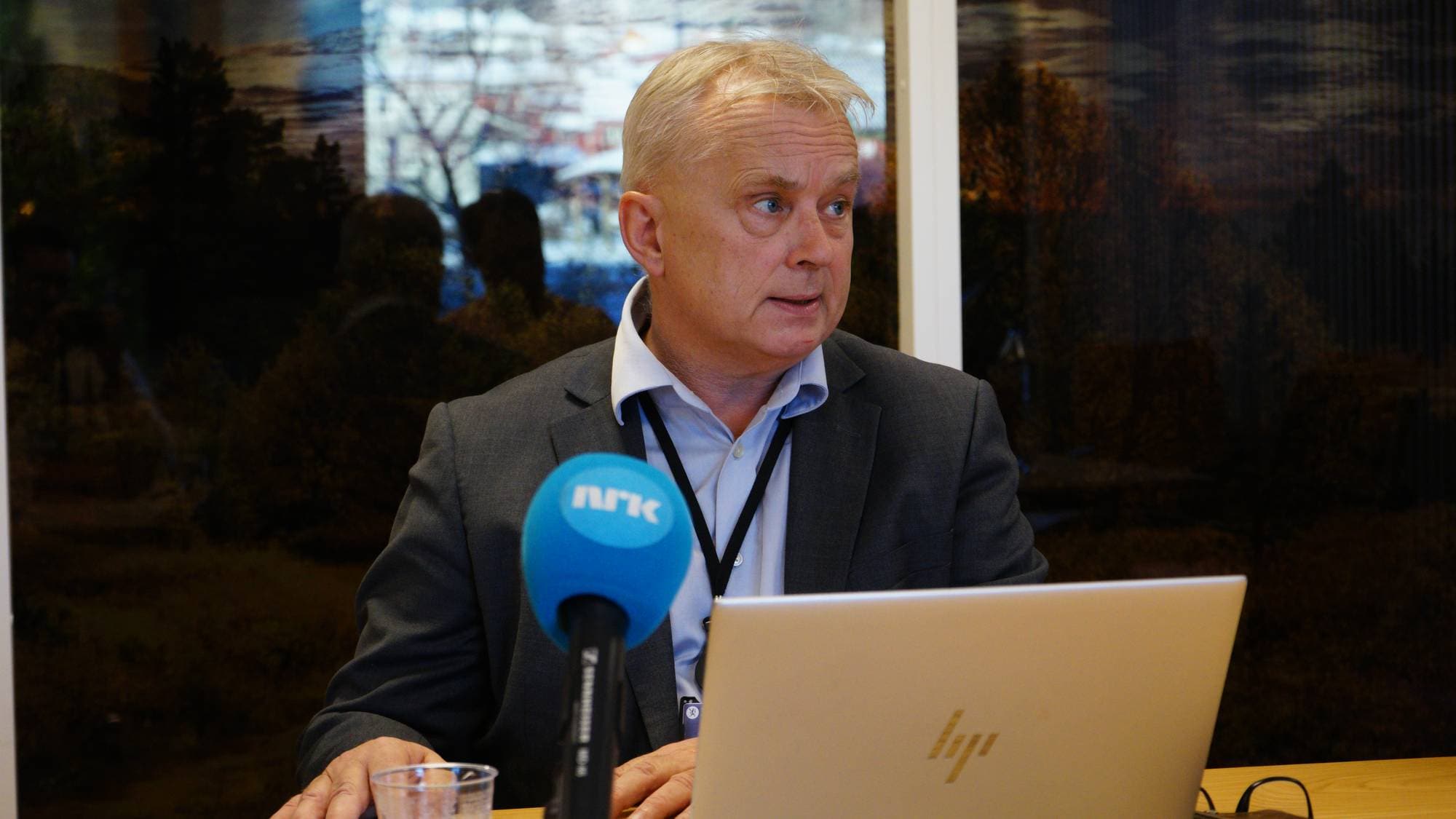 En mann sitter foran en laptop ved et bord, med en mikrofon og et glass vann foran seg. Han har på seg en grå jakke og blå skjorte, mens en svart lanyard henger rundt halsen hans. I bakgrunnen vises en utsikt over en naturscene. Rommet er lyst, med vinduer som slipper inn naturlig lys.