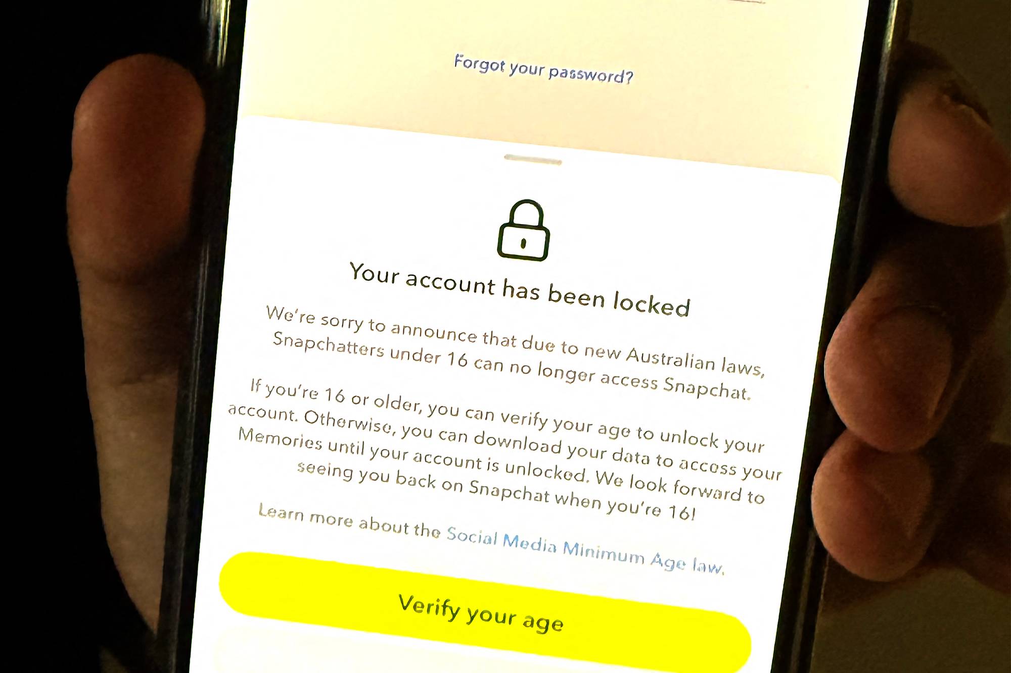 En mobil viser et skjermbilde med en tydelig Snapchat-gul knapp merket "Verify your age". Budskapet på skjermen er at Snapchat ikke lenger er tilgjengelig i Australia for brukere under 16 år.