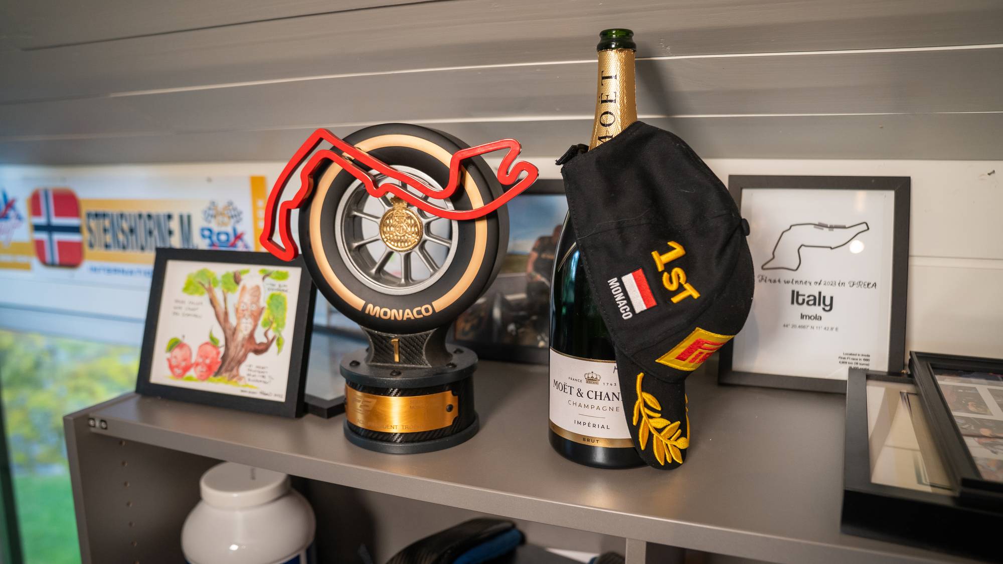 Pokalen for Monaco, en flaske champagne og en caps ligger på en hylle. Capsen har "1st" og Monacos flagg. Til venstre er et bilde av et tre og epler. Til høyre er et bilde av en racerbane i Imola. (Bildebeskrivelsen er laget av en KI-tjeneste)