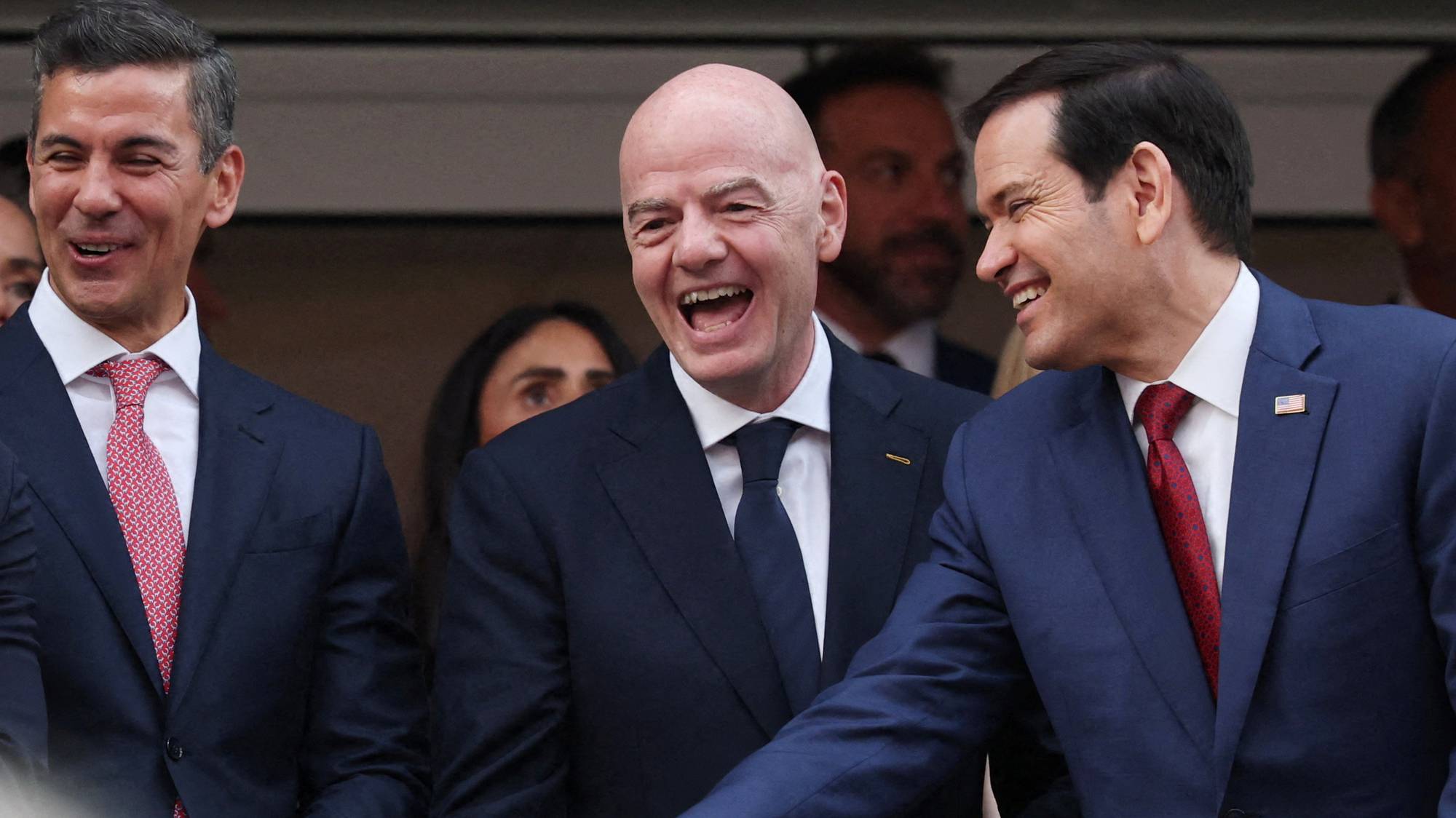 Gianni Infantino og Marco Rubio ler sammen med andre menn i dress på en tribune. (Bildebeskrivelsen er laget av en KI-tjeneste)