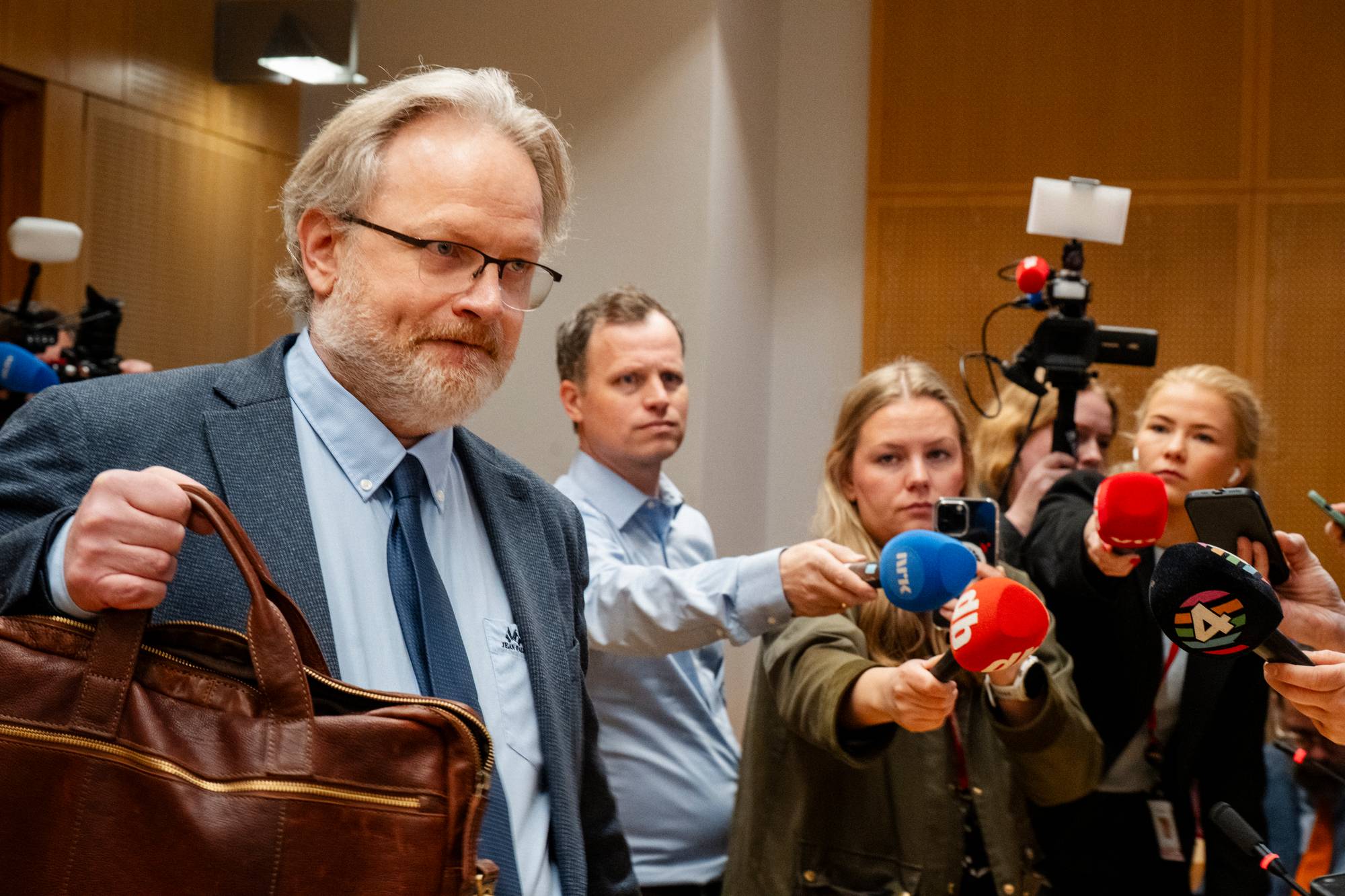 En mann i dress med dokumentveske går forbi en gruppe journalister med mikrofoner. Journalister holder opp mikrofoner og telefoner mot mannen. Det er et kamera på stativ bak dem. (Bildebeskrivelsen er laget av en KI-tjeneste)